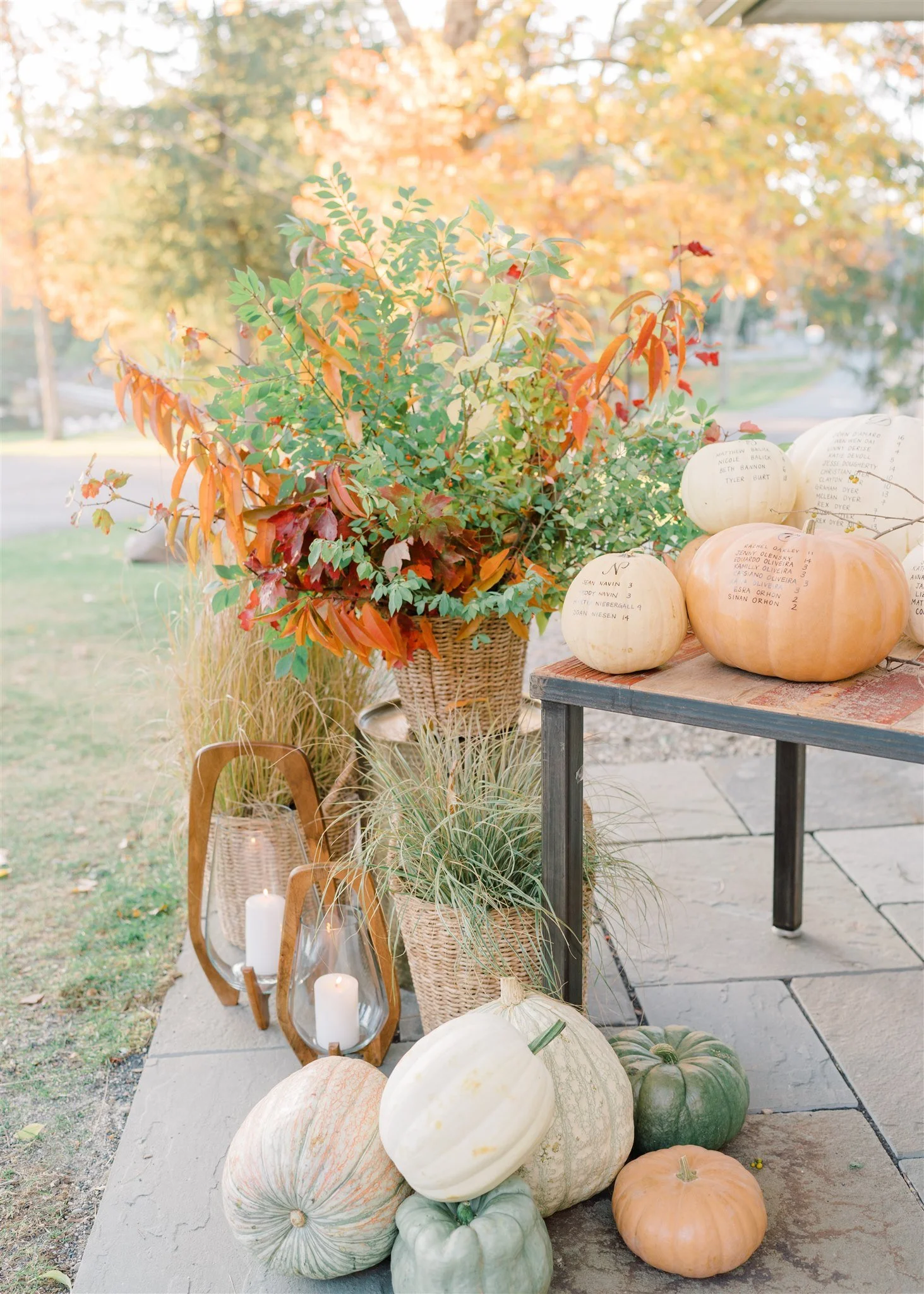 Decorative fall display with white, green, and orange pumpkins, potted plants, candles, and a wicker basket outdoors on a stone patio, with trees in the background.