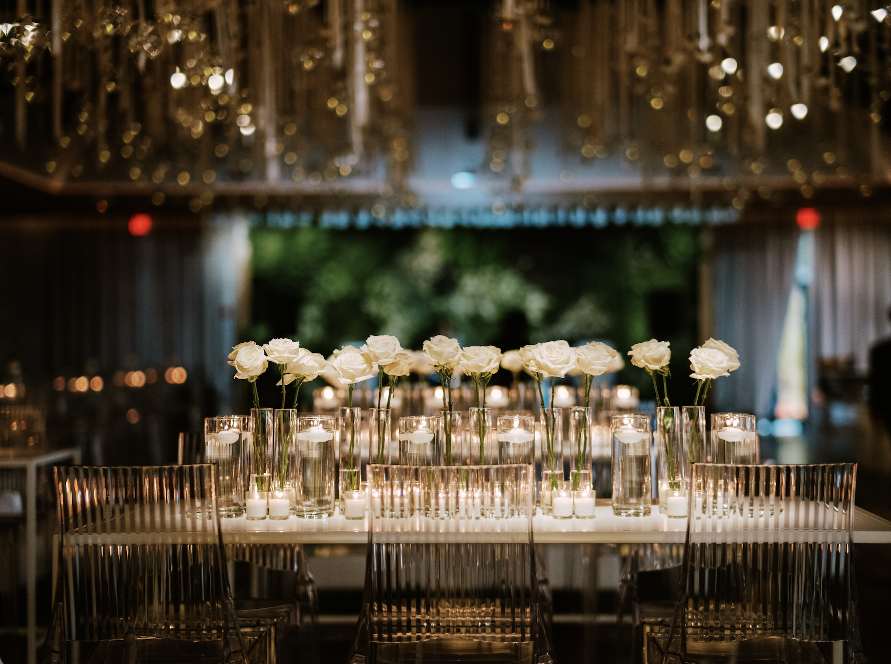 Elegant dinner table decorated with white roses in tall glass vases and floating candles in small glass holders, set up in an indoor event space with ambient lighting, greenery outside visible through open curtains.