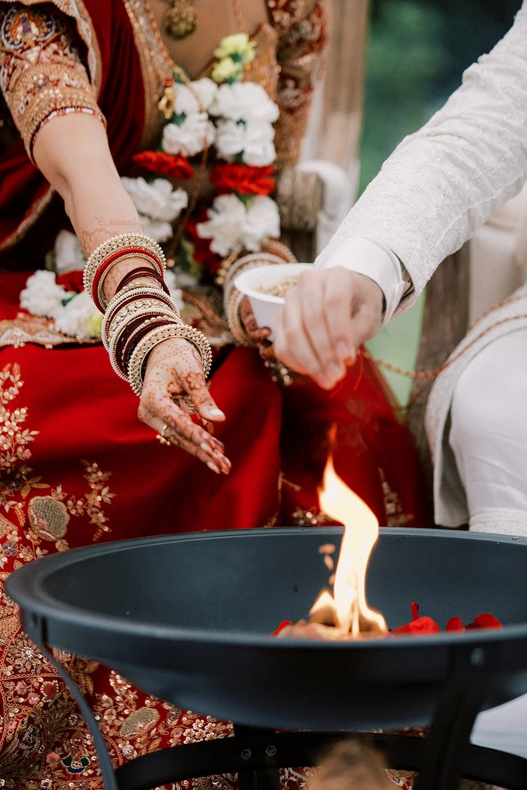 Indian bride and groom performing a traditional wedding ritual with fire, wearing elaborate attire and jewelry, with flower garlands around their necks.