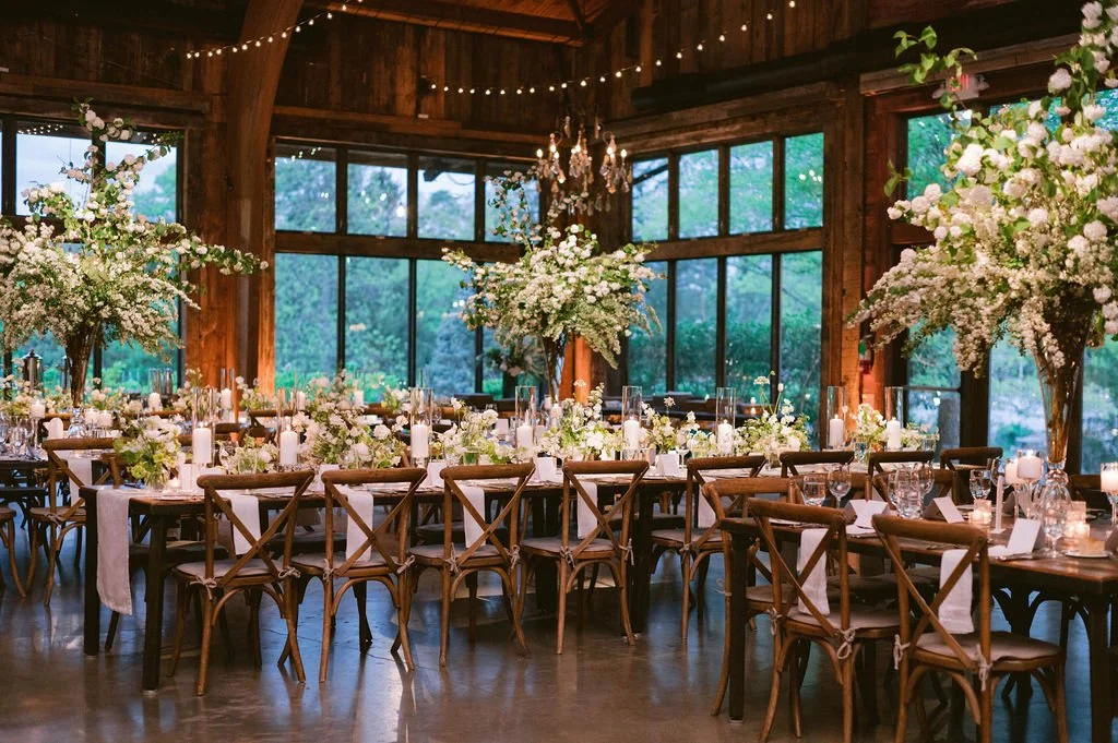 An indoor wedding reception setup with long tables decorated with white floral centerpieces, candles, and glassware. The room features large windows, wooden walls, and string lights hanging from the ceiling, creating a warm and elegant atmosphere.