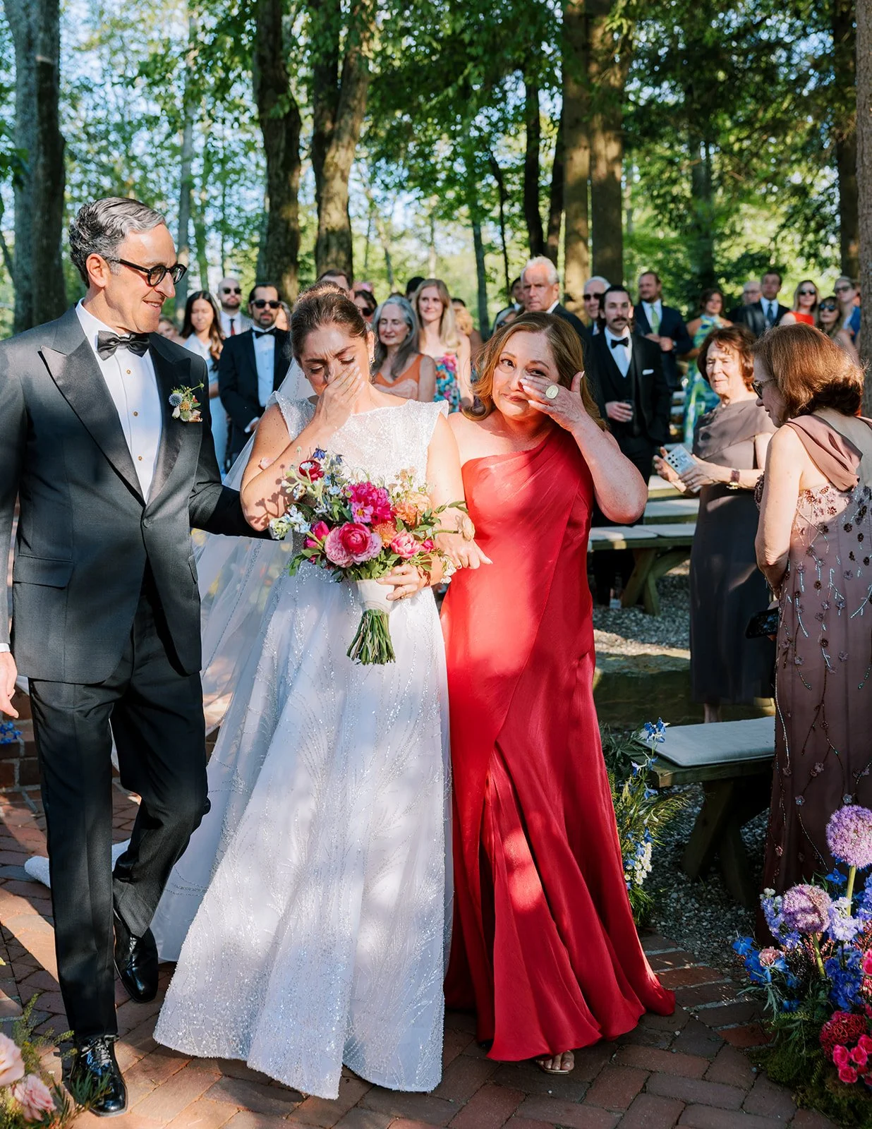 A bride in a white gown holding a bouquet of pink and purple flowers, emotional during her wedding ceremony outdoors surrounded by family and friends.