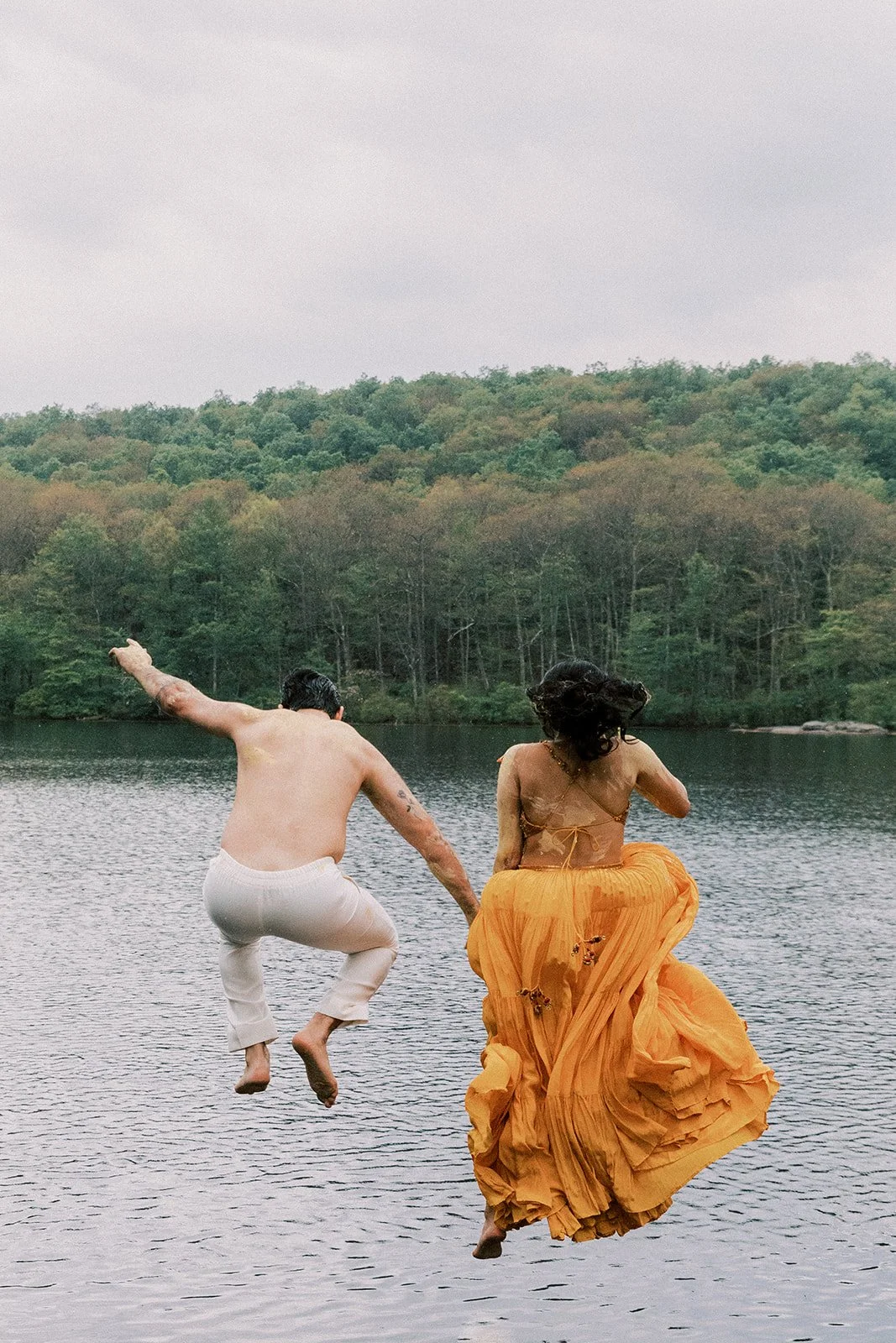 A man and woman jumping into a lake, with the man wearing white pants and no shirt, and the woman wearing a flowing orange skirt and top, against a background of trees and a cloudy sky.