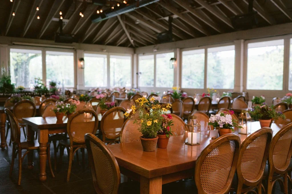 A decorated indoor event space with wooden tables, floral centerpieces, candles, and large windows showing greenery outside.