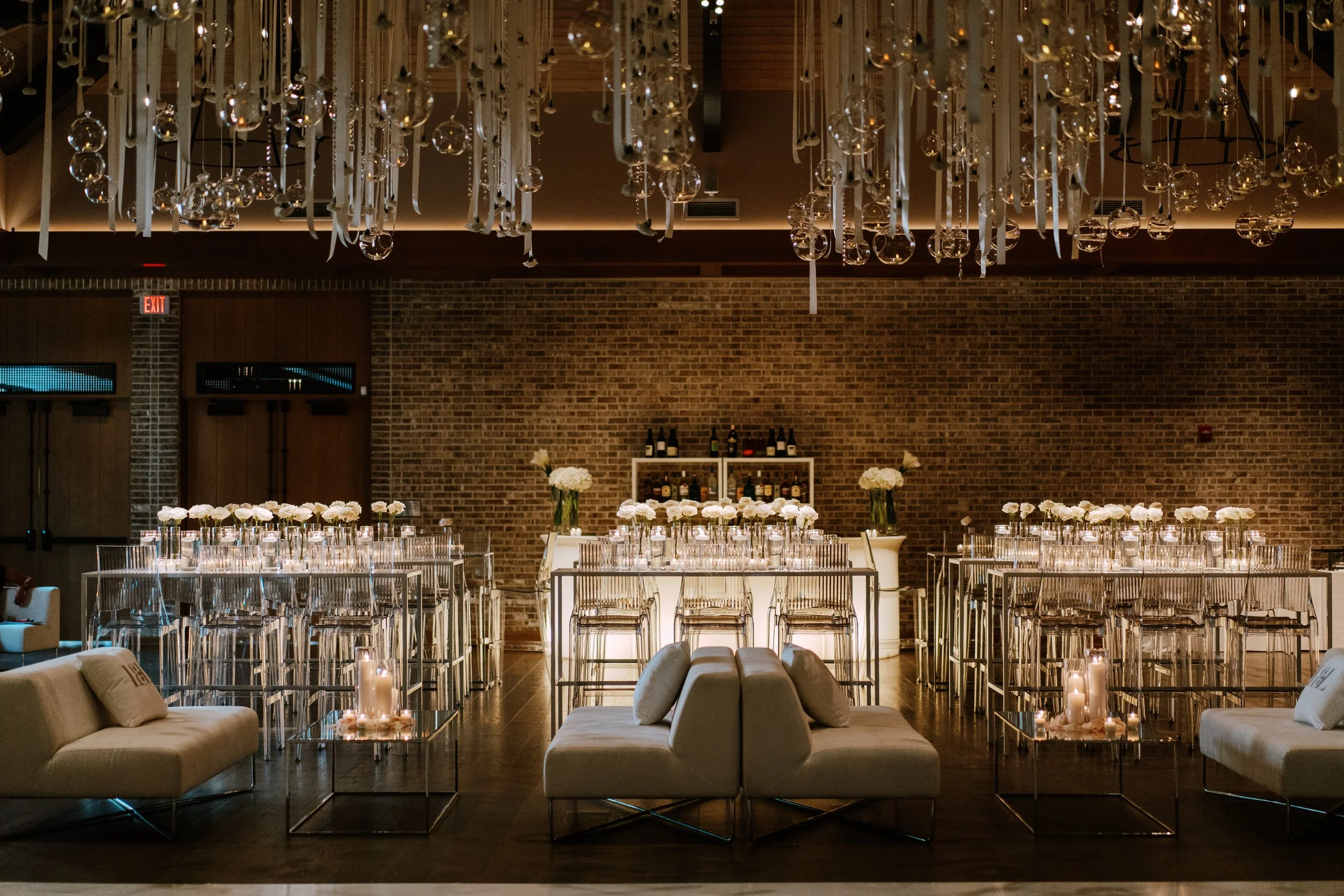 Elegant event space with a brick wall background, decorated with white flowers in vases, candles on glass tables, modern white sofas, and a chandelier with hanging ornaments.