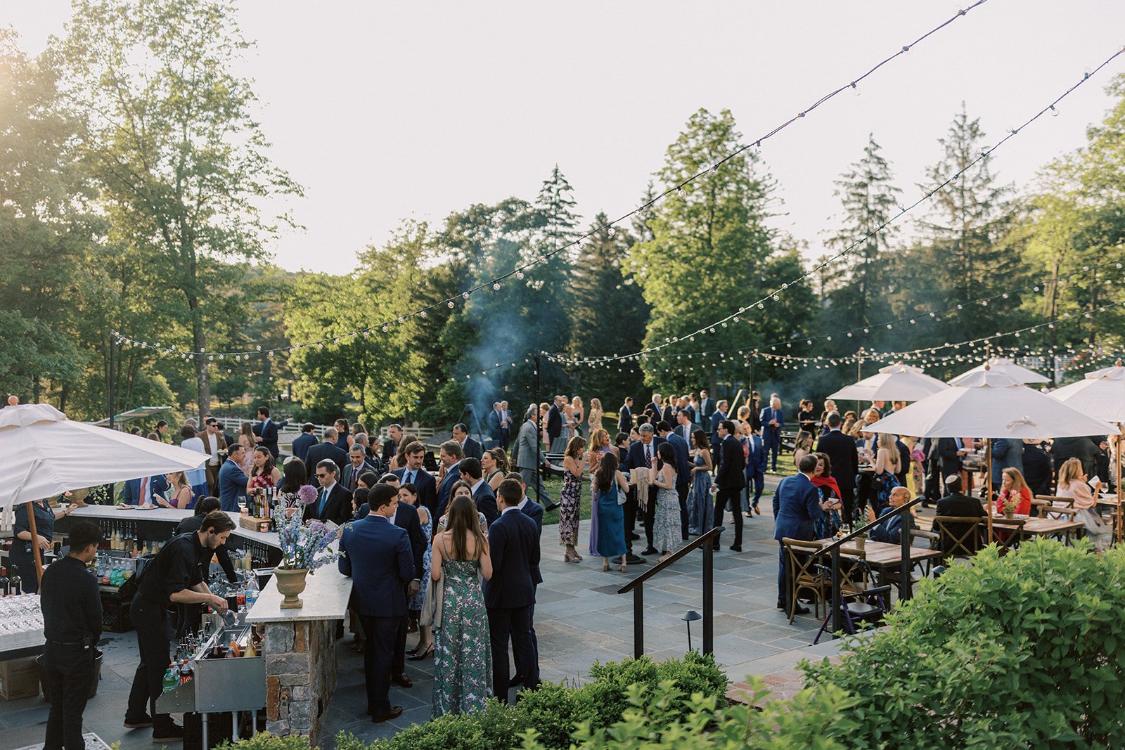 A large outdoor social gathering with people dressed in formal attire, mingling under string lights on a patio surrounded by trees, with umbrellas and a bar area.
