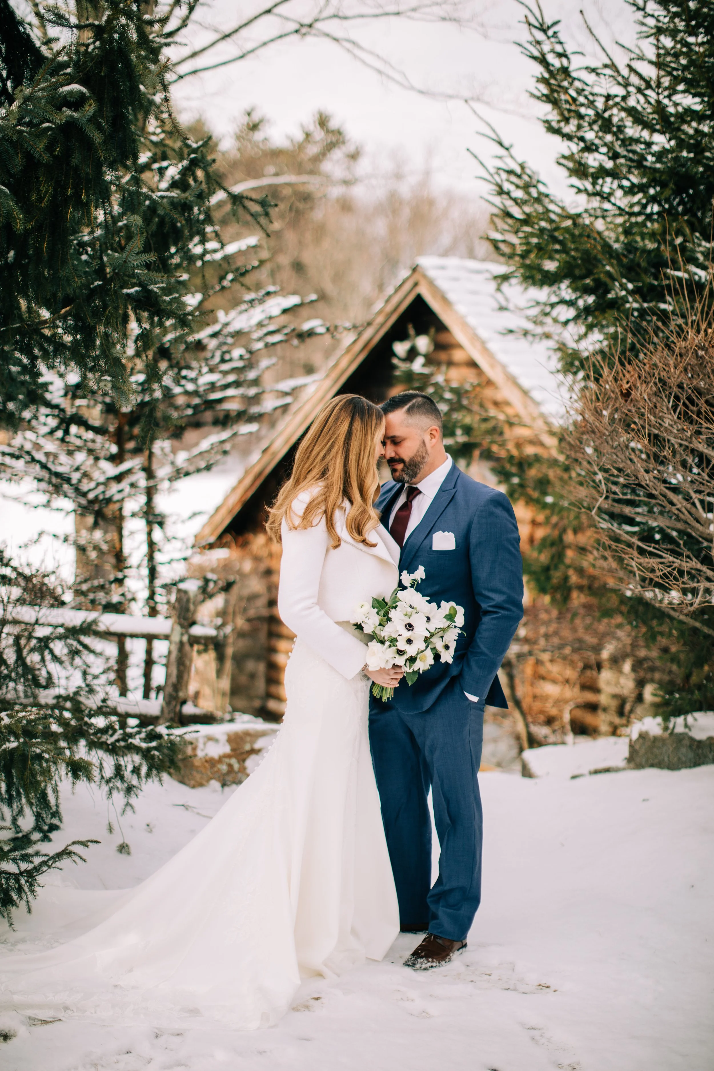 A bride and groom standing closely together outdoors in a snowy landscape, with the bride holding a bouquet of white flowers, near a rustic wooden cabin and surrounded by evergreen trees.