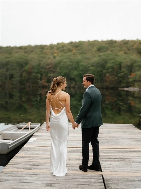 A bride and groom holding hands on a wooden dock by a lake, with trees in the background, during daytime.
