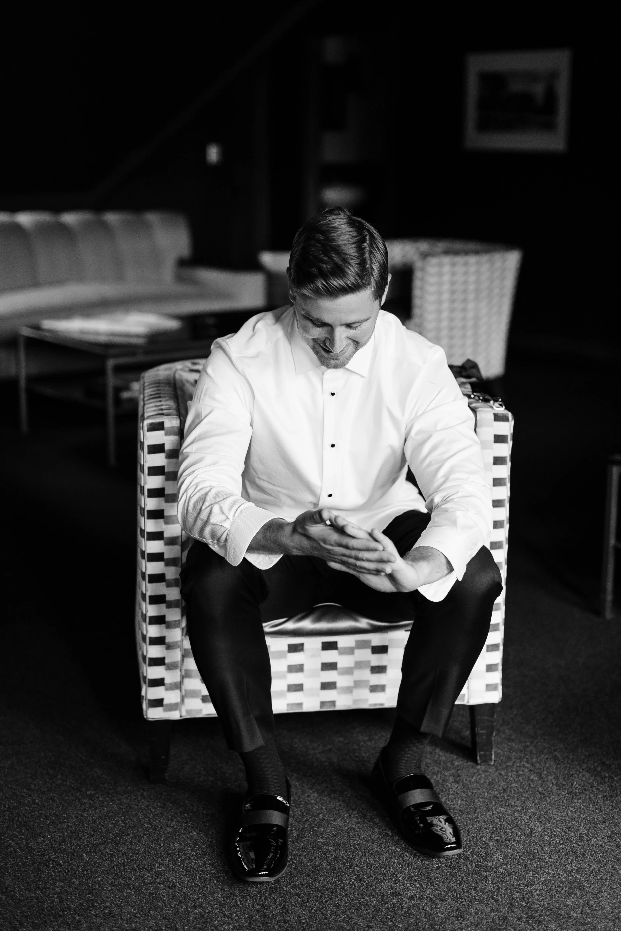 A man in a white dress shirt and black pants sitting on a patterned armchair, smiling and looking down, in a dimly lit room with furniture and framed pictures in the background.