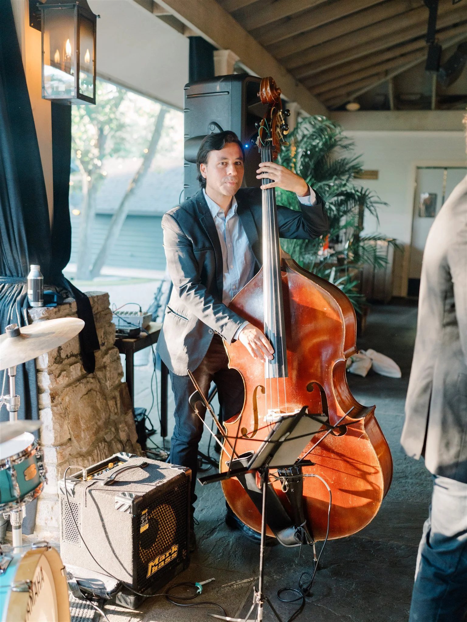 A man wearing a blazer playing a double bass during a live music performance in a cozy indoor setting with large windows and green plants.