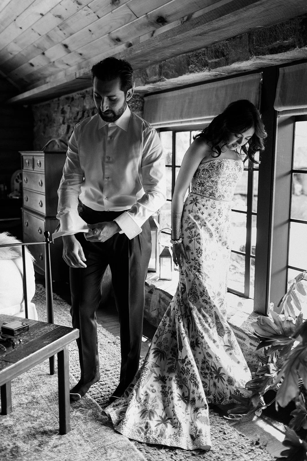 A man and a woman dressed formally inside a rustic room, with large windows and a wooden ceiling, as the man adjusts his cufflink.