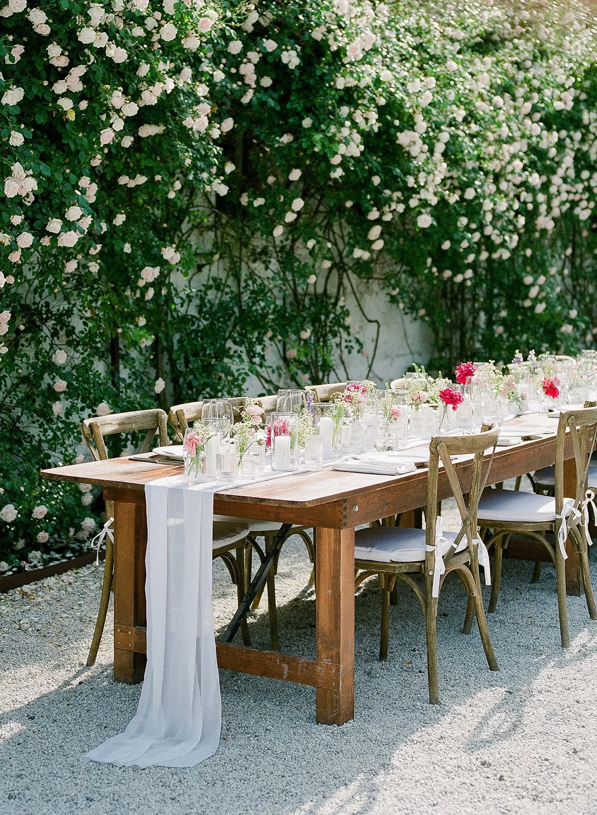 A long wooden table set for outdoor dining with white table runner, glassware, white plates, and pink floral centerpieces, surrounded by wooden chairs with white cushions, in front of a backdrop of green foliage and white flowering bushes.