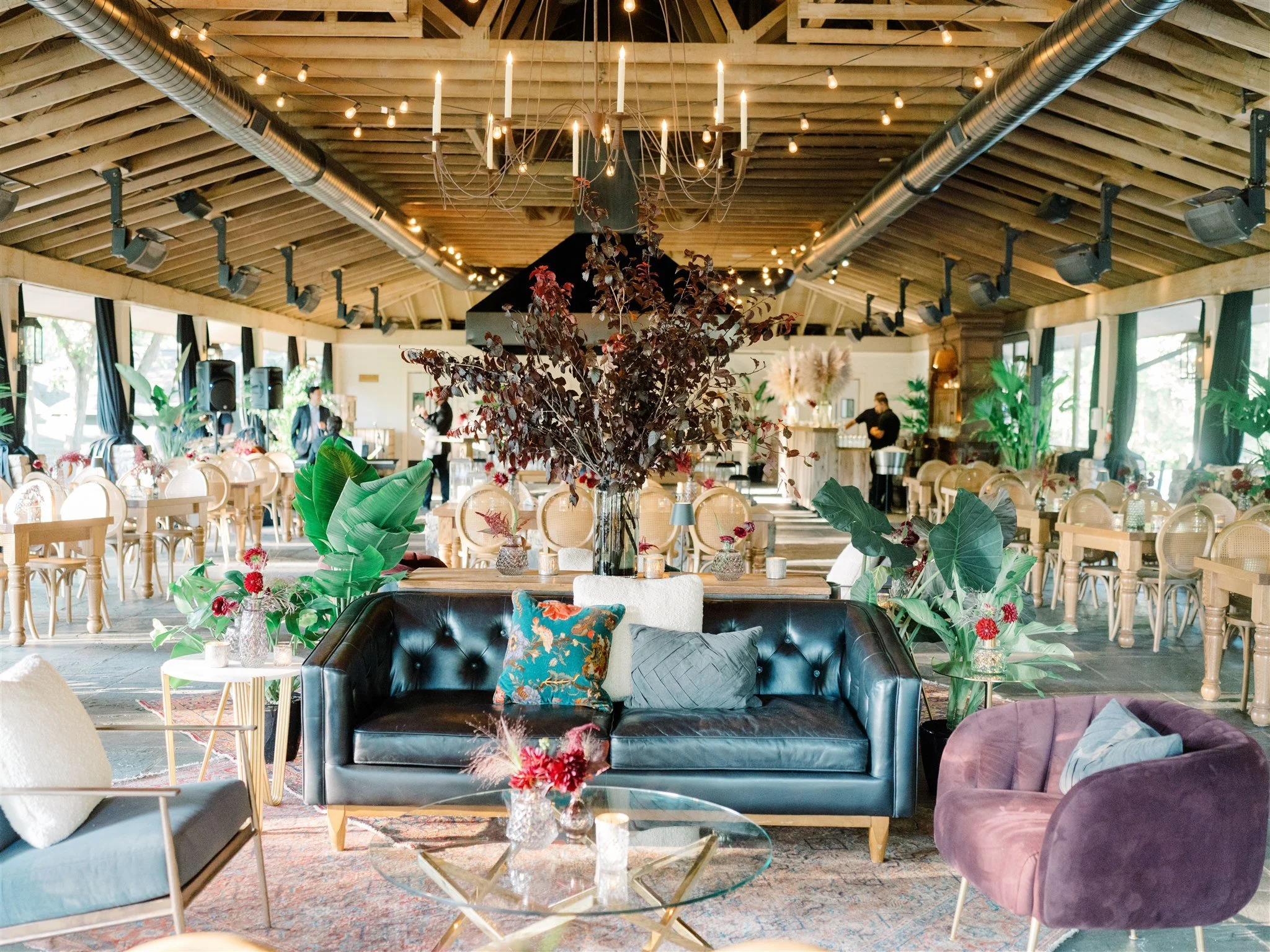 Interior of a decorated event space with a black leather sofa, purple velvet chair, glass coffee table, and floral arrangements, with wooden tables and chairs, string lights, and large windows.