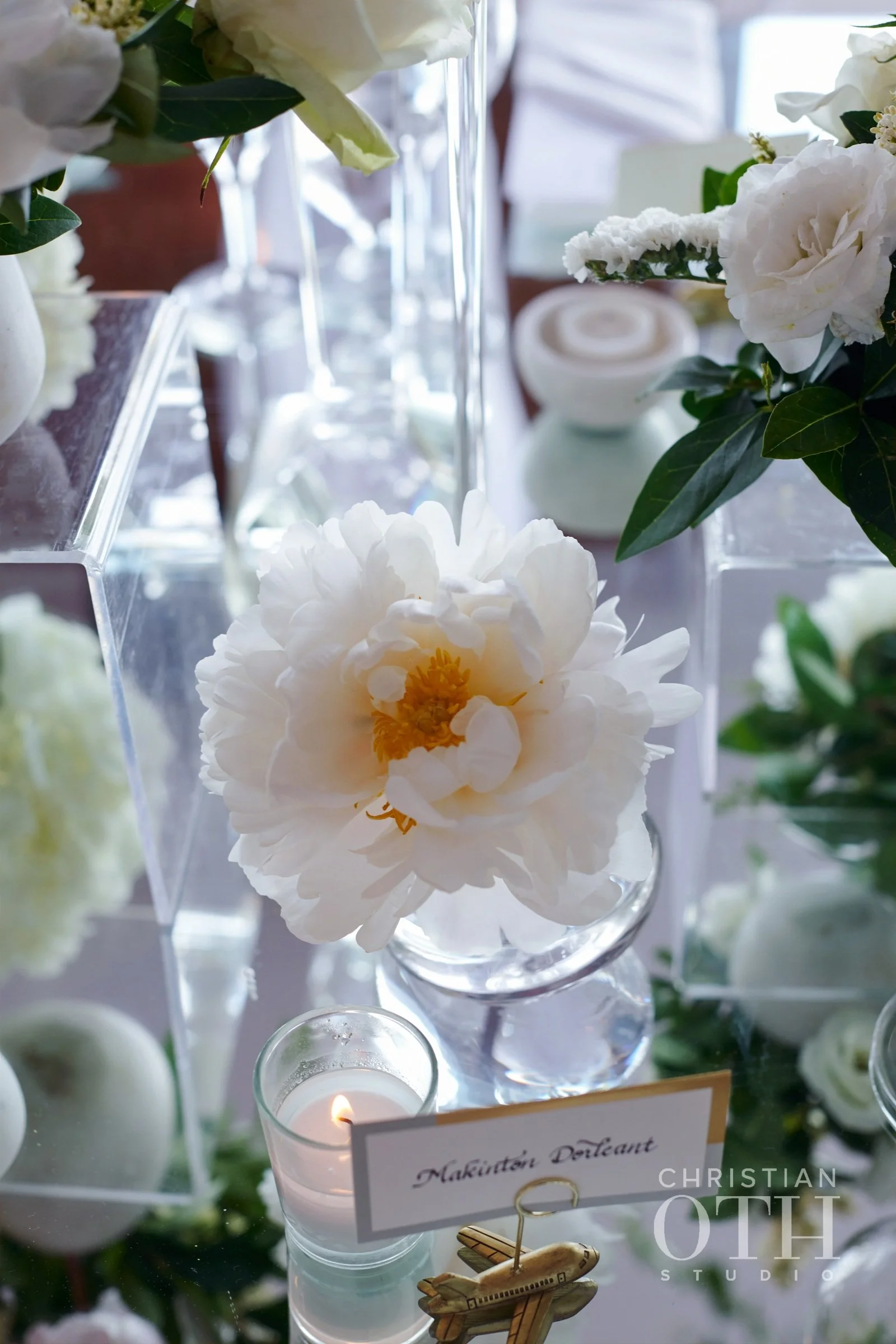 A close-up of white peony flowers in a glass vase on a table, with a lit candle in a glass holder and a small airplane-shaped decoration nearby, at a floral arrangement display or event.
