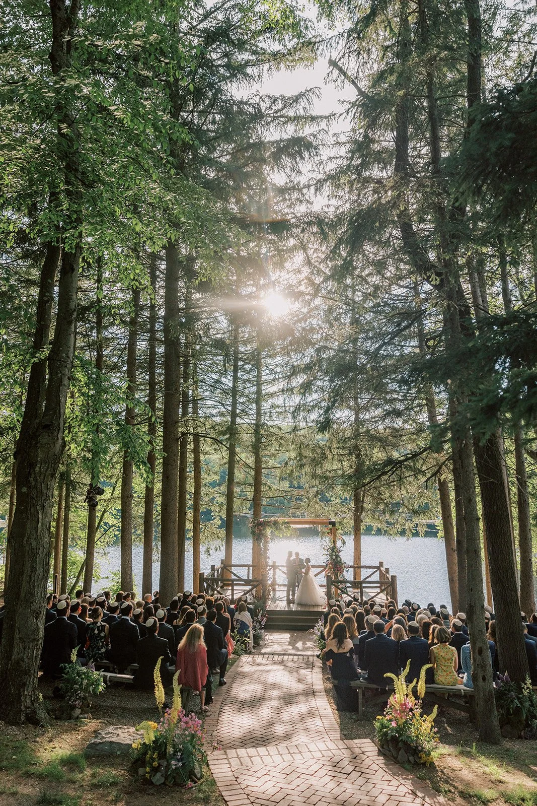 Outdoor wedding ceremony by a lake, surrounded by tall trees with the sun shining through, guests seated on benches, and a couple at the altar.