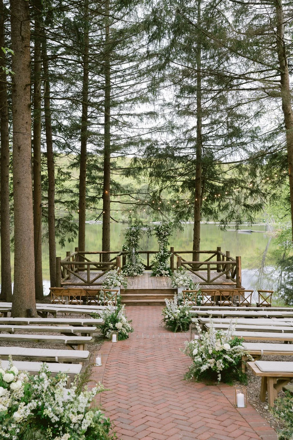 Outdoor wedding ceremony setup with benches, floral arrangements, a small wooden platform, and string lights in a wooded area by a lake.