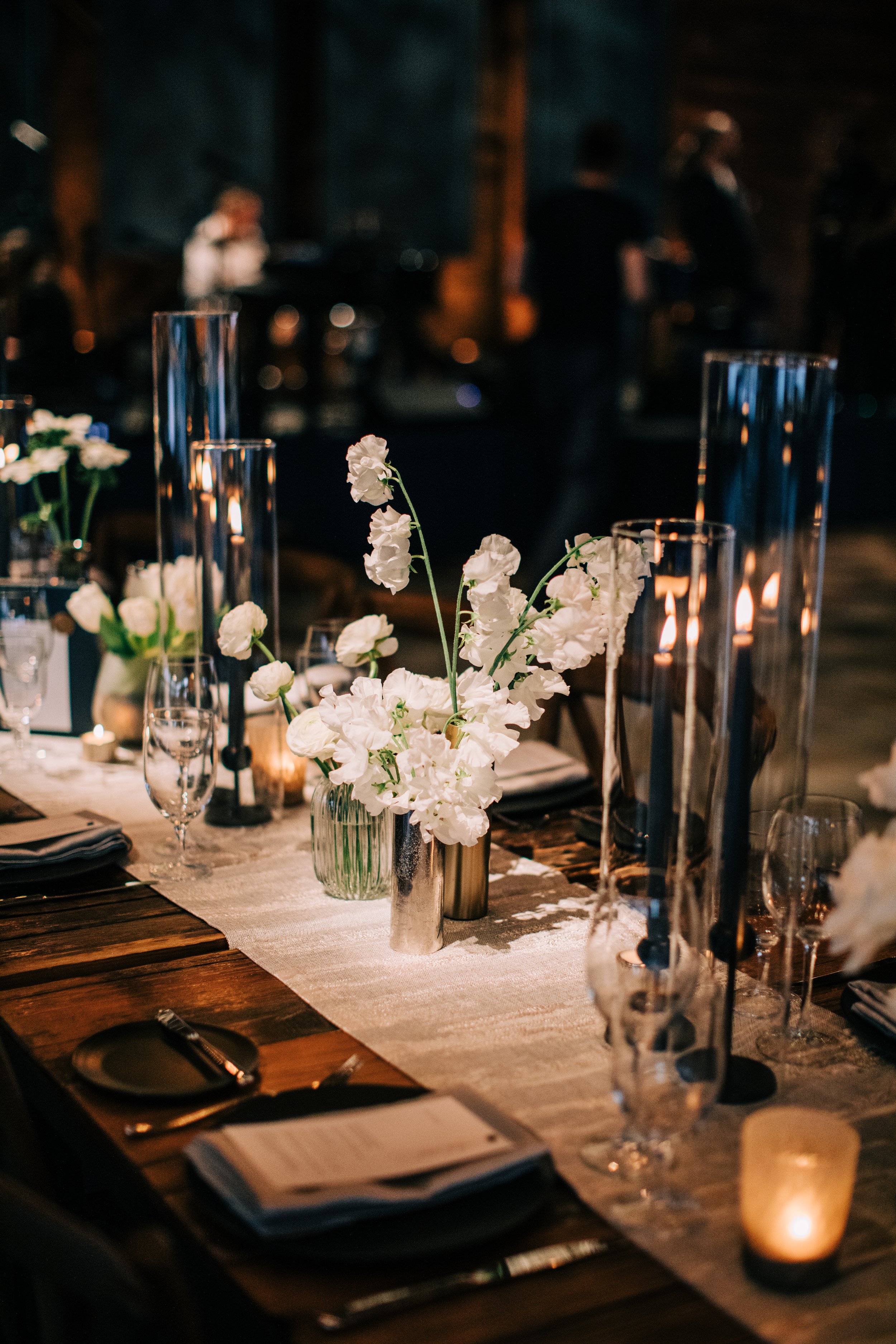 A decorated table with white flowers in vases, lit candles, glassware, and place settings, set for an event or dinner in a dimly lit room.