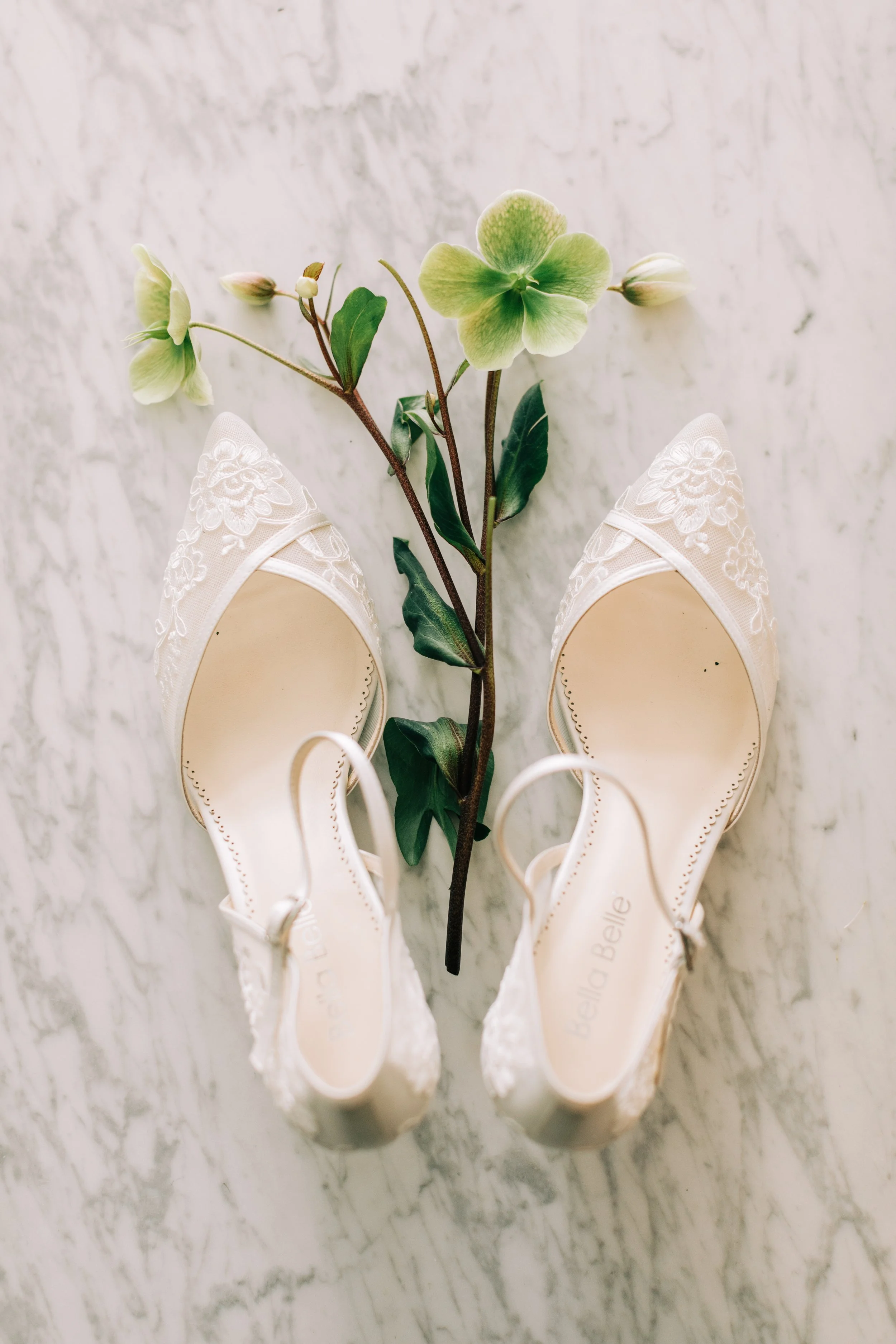 A pair of white, embroidered wedding shoes with floral designs, placed on a marble surface, with a green flower lying between them.