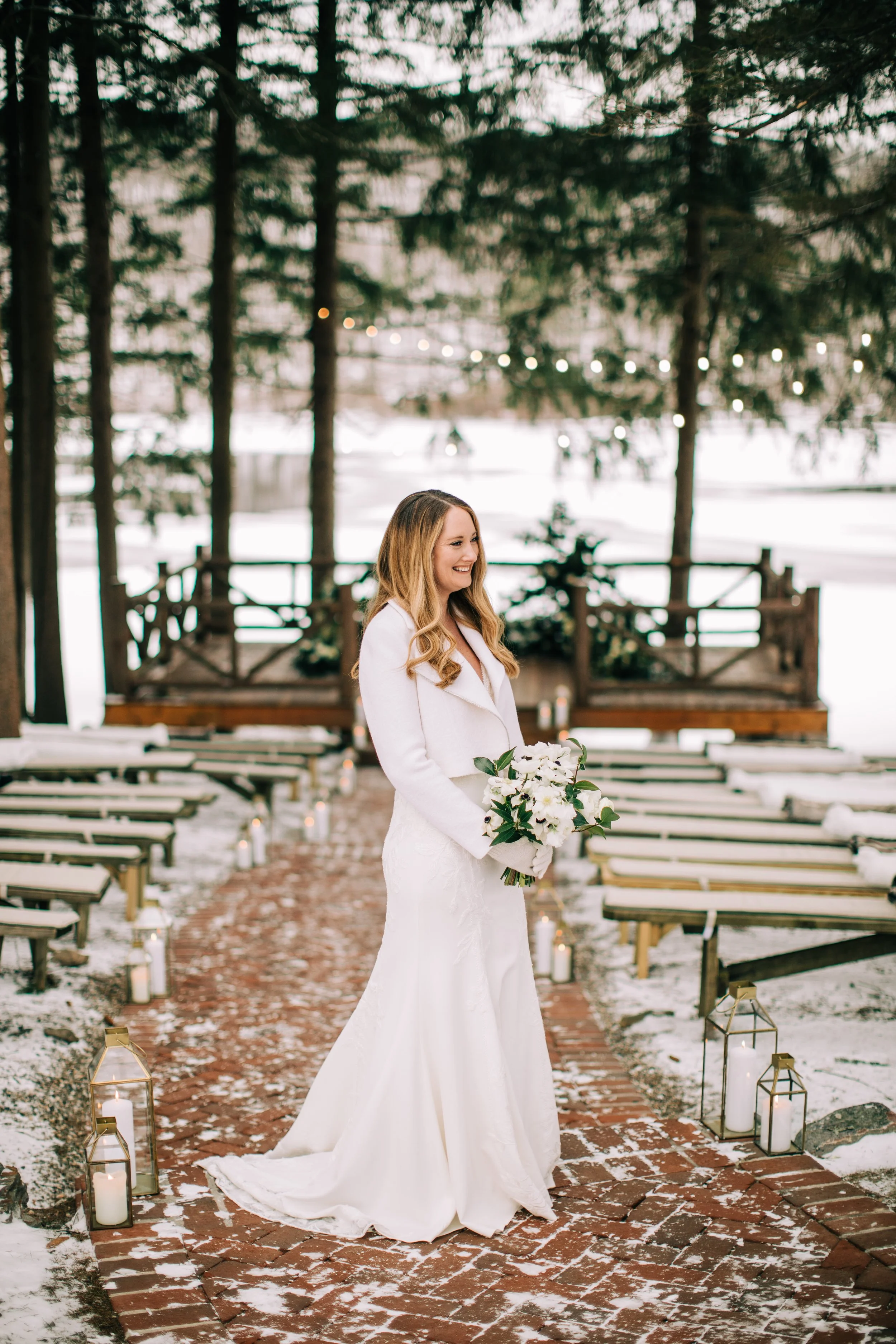 A bride in a white wedding dress holding a bouquet of white flowers, standing on a brick pathway outdoors with snow, surrounded by lanterns and trees.