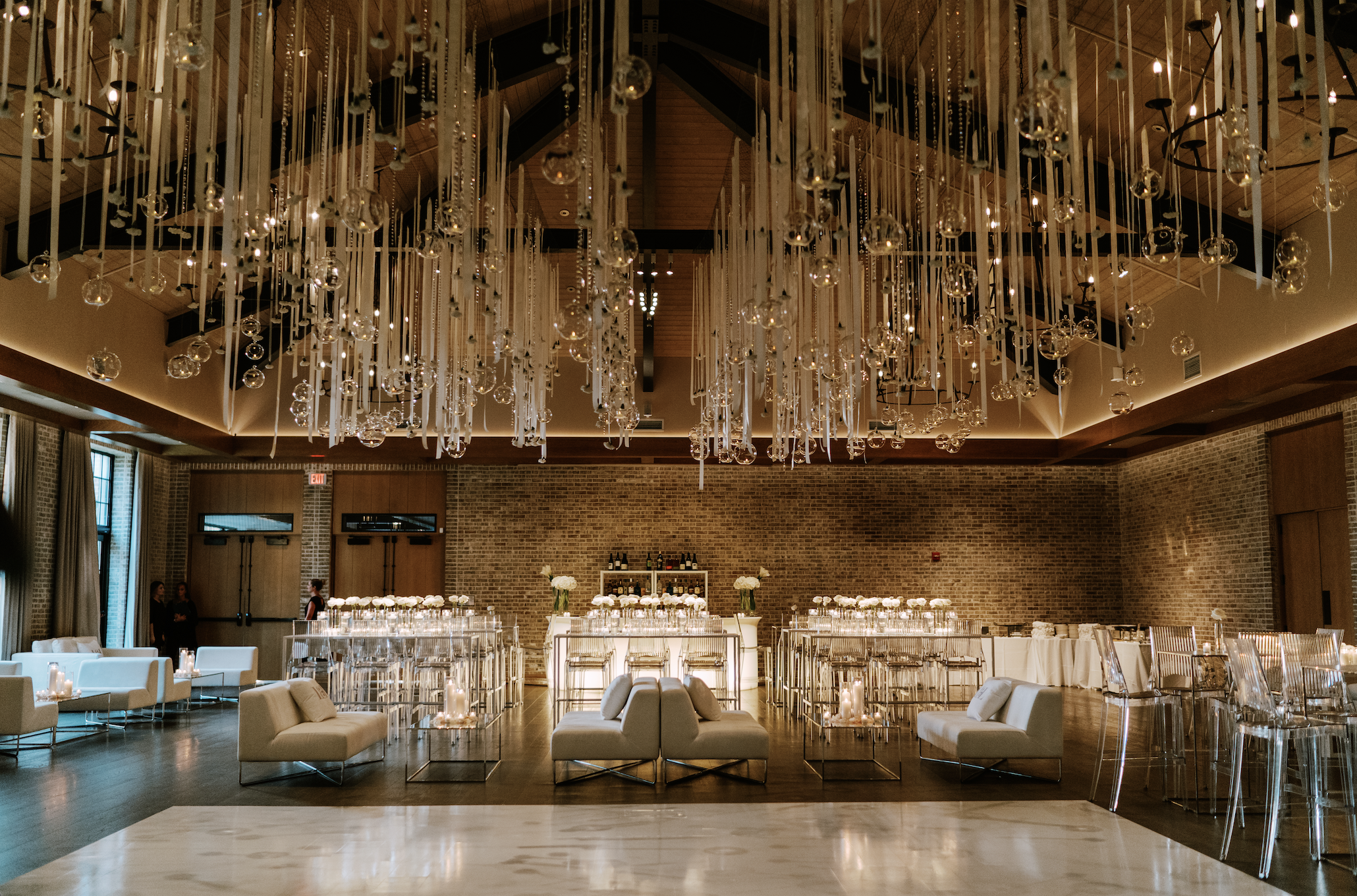 Elegant event space with hanging glass orbs and ribbons, white couches with cushions, glass tables with candles, bar area with bottles and flowers, wooden floors, brick wall, large windows, and ambient lighting.