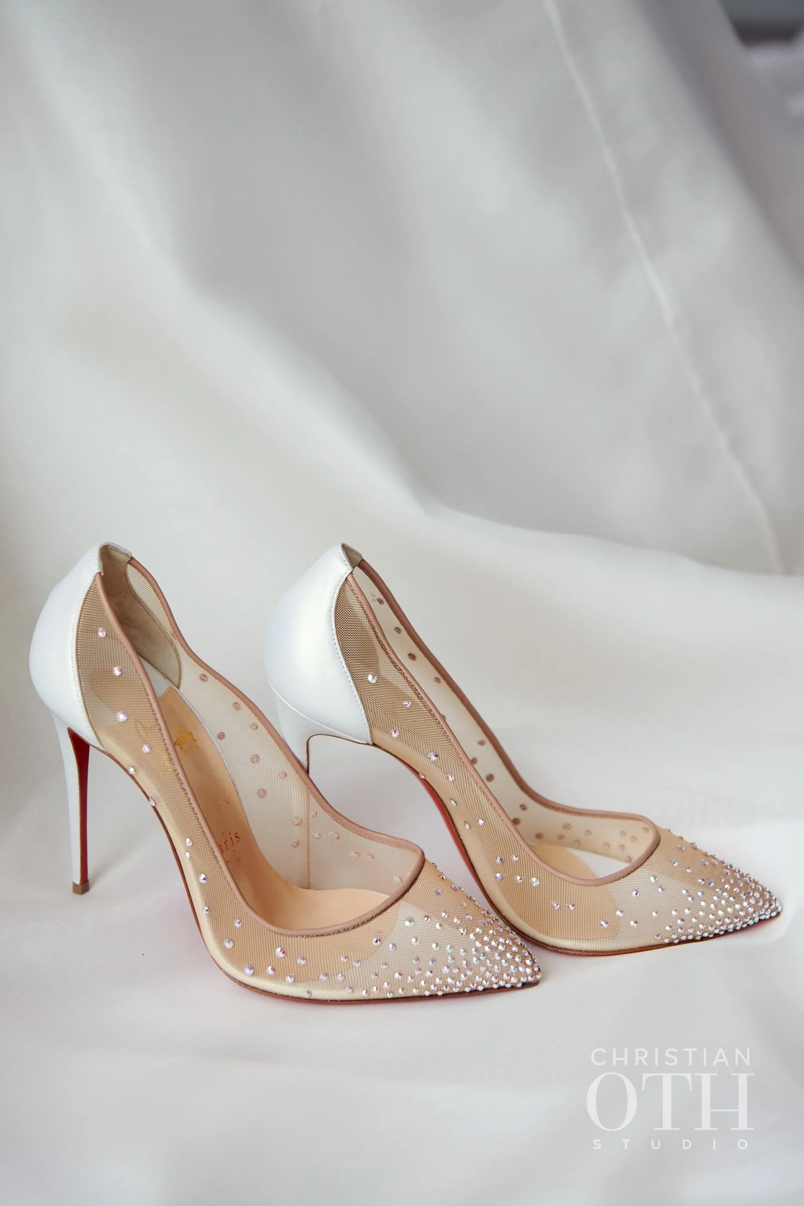 Pair of elegant high-heeled shoes with pointed toes, beige and white design, adorned with small sparkling rhinestones, displayed on a white surface with a soft background.