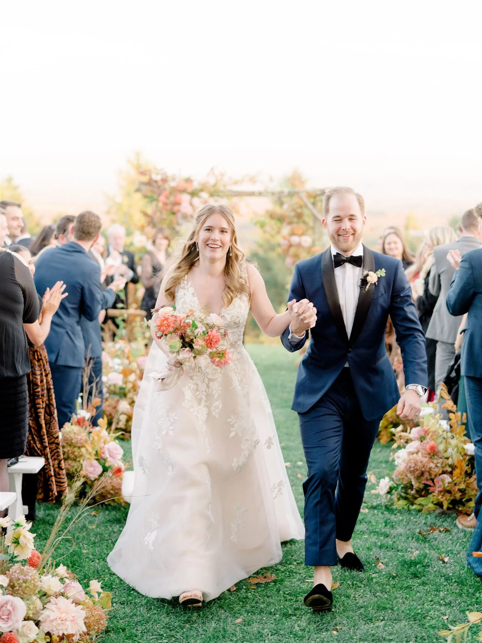 A newlywed couple walks hand-in-hand down the aisle during an outdoor wedding ceremony, surrounded by friends and family, with floral decorations and trees in the background.