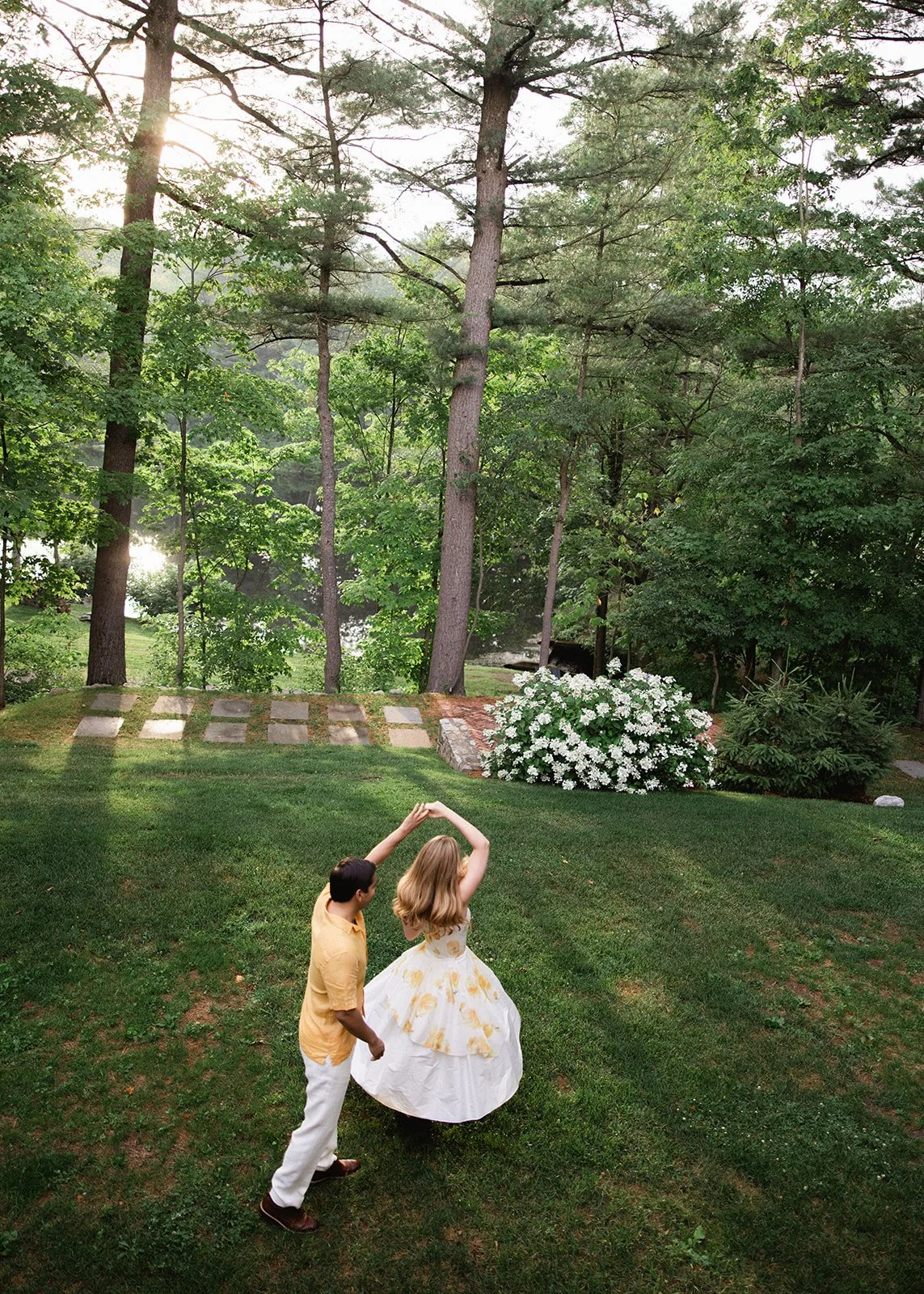 A young man and woman dancing outdoors in a lush green garden surrounded by tall trees and blooming white flowers.