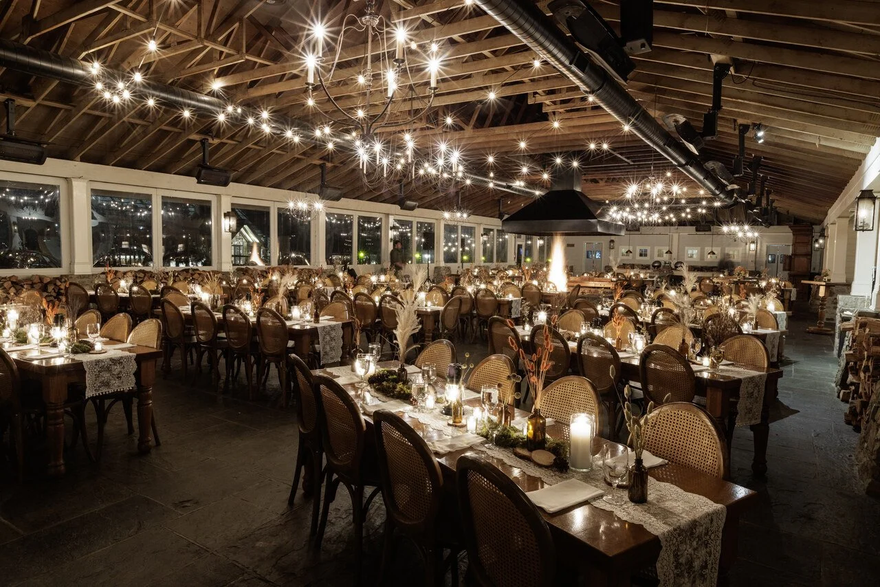 A decorated banquet hall with long tables set with candles, vases, and tableware, under a vaulted wooden ceiling with string lights and chandeliers, prepared for an event or gathering.
