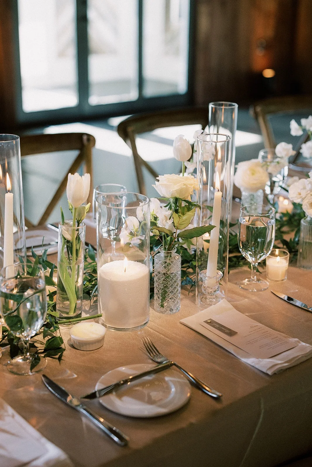 Elegant dining table set with white flowers, candles, water glasses, a menu, and silverware in a warmly lit room.