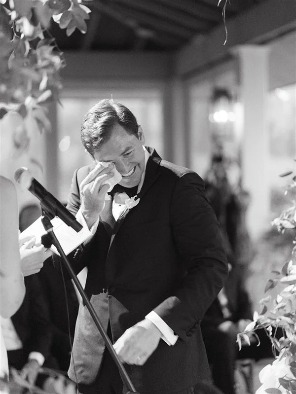Man in a tuxedo crying tears of joy at what appears to be a wedding reception.