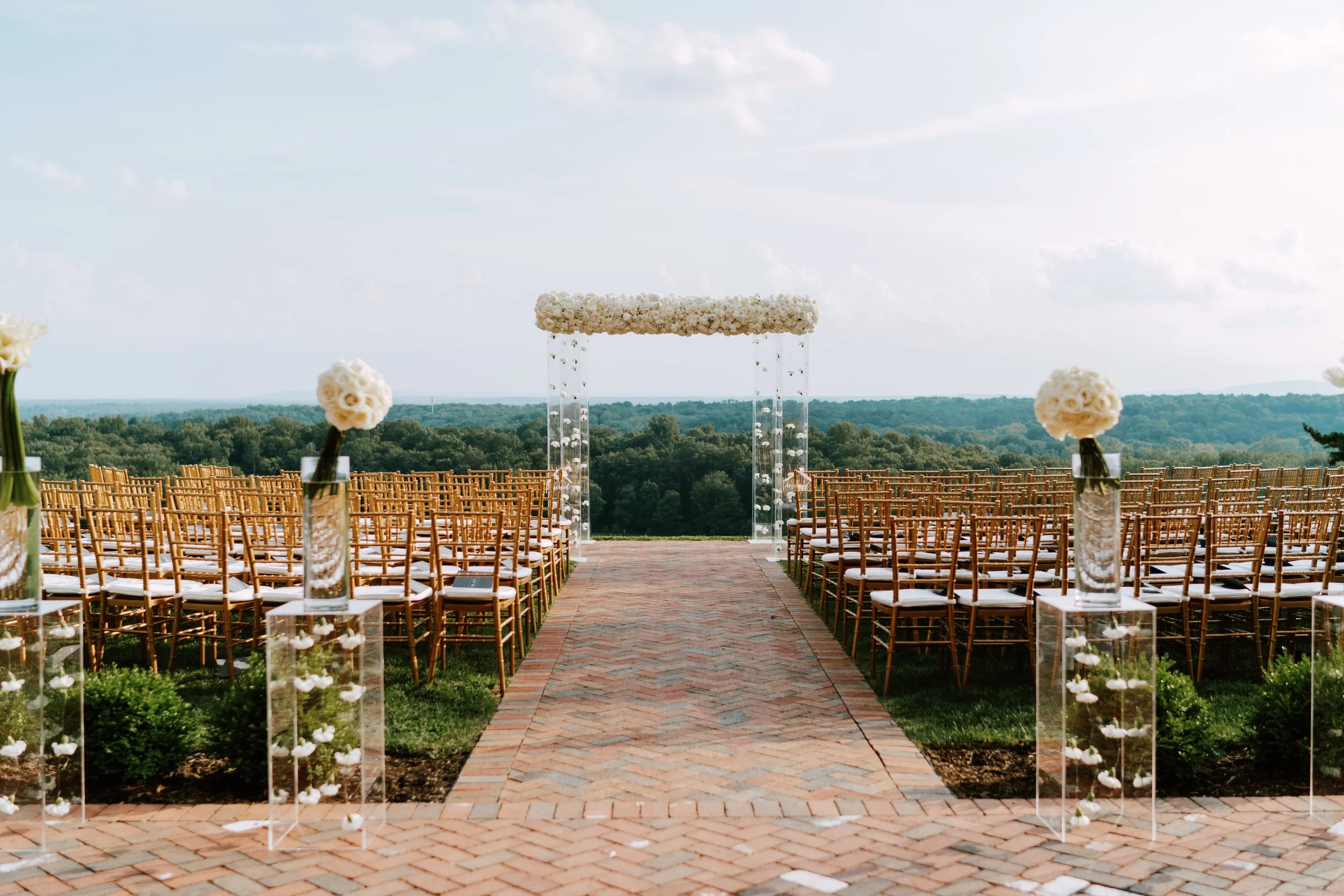 Outdoor wedding ceremony setup with rows of wooden chairs facing a floral arch, clear vases with white flowers, on a brick pathway overlooking a landscape with trees and a cloudy sky.