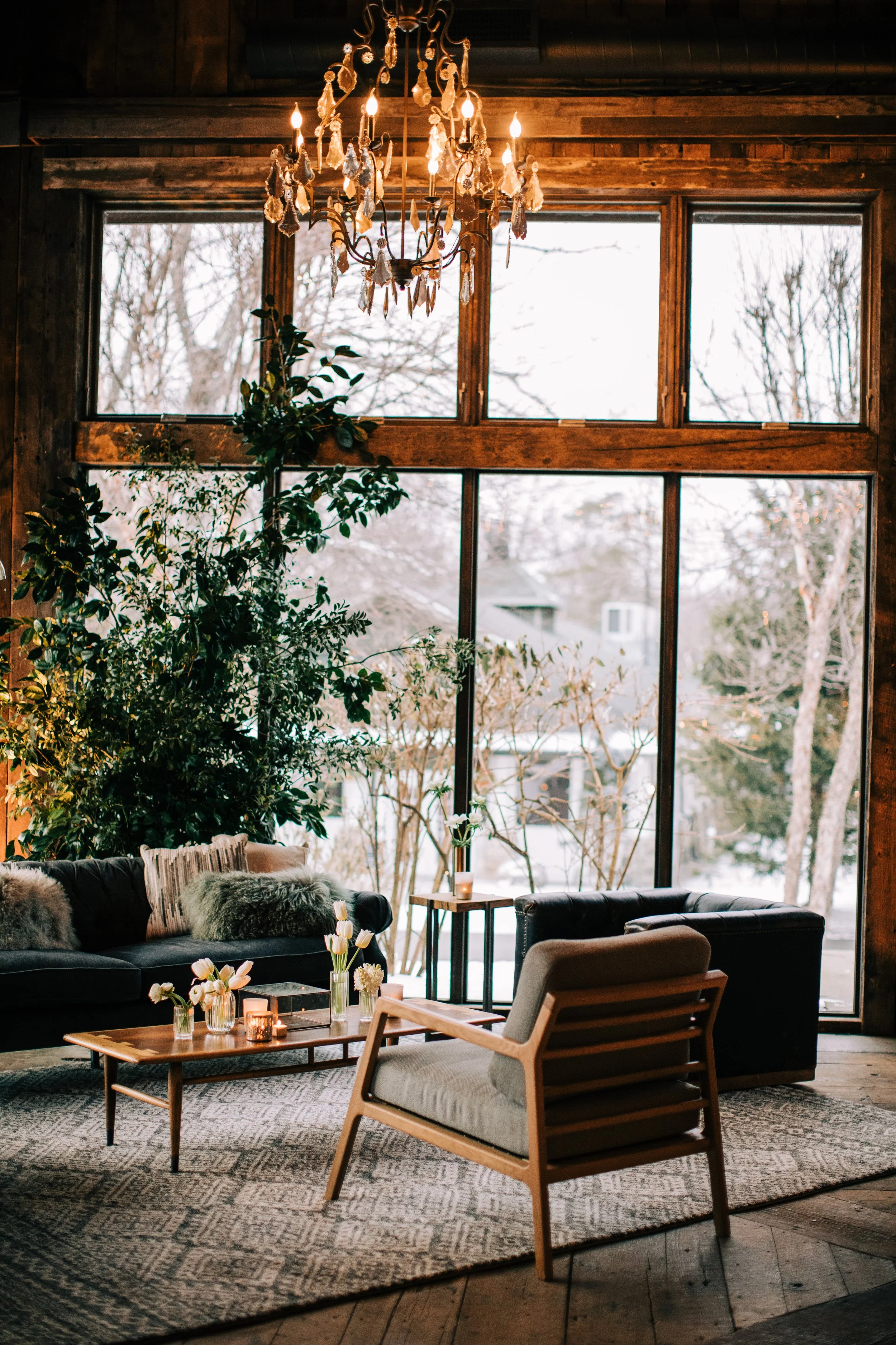 Cozy living room with large windows, wooden accents, a chandelier, plush pillows, a modern armchair, a leather sofa, a patterned rug, and a wooden coffee table decorated with vases of flowers and candles.