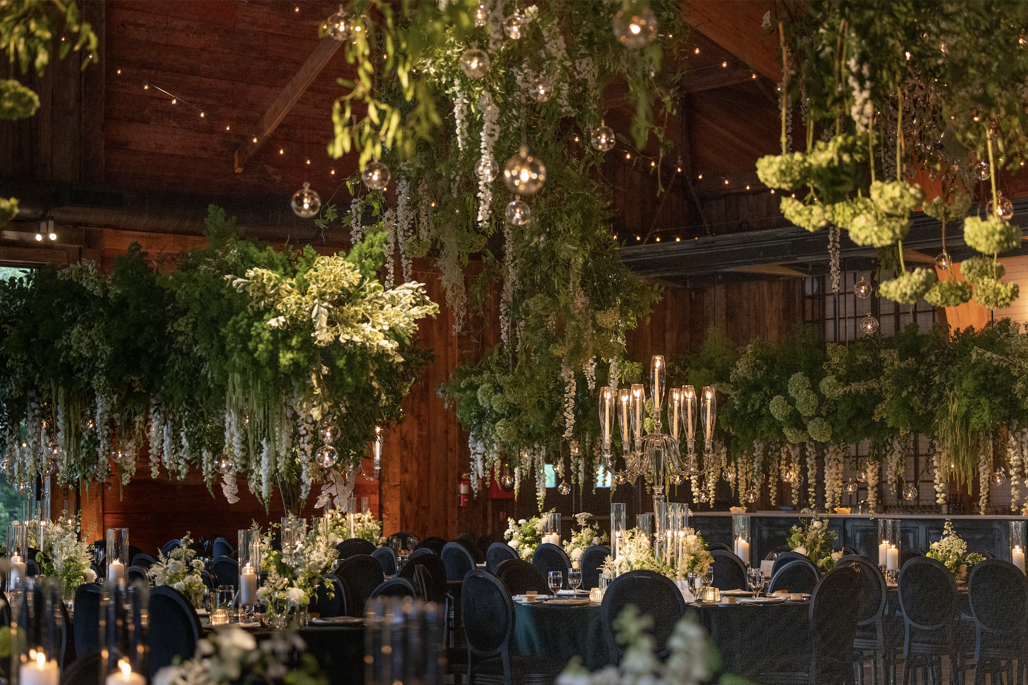Elegant event space decorated with hanging greenery and white flowers, candle centerpieces, and string lights, with dark chairs around tables.