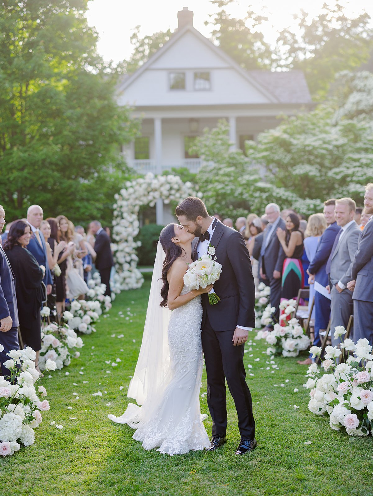 A newly married couple kisses at their outdoor wedding ceremony as guests look on, with a white floral arch and a large white house in the background.