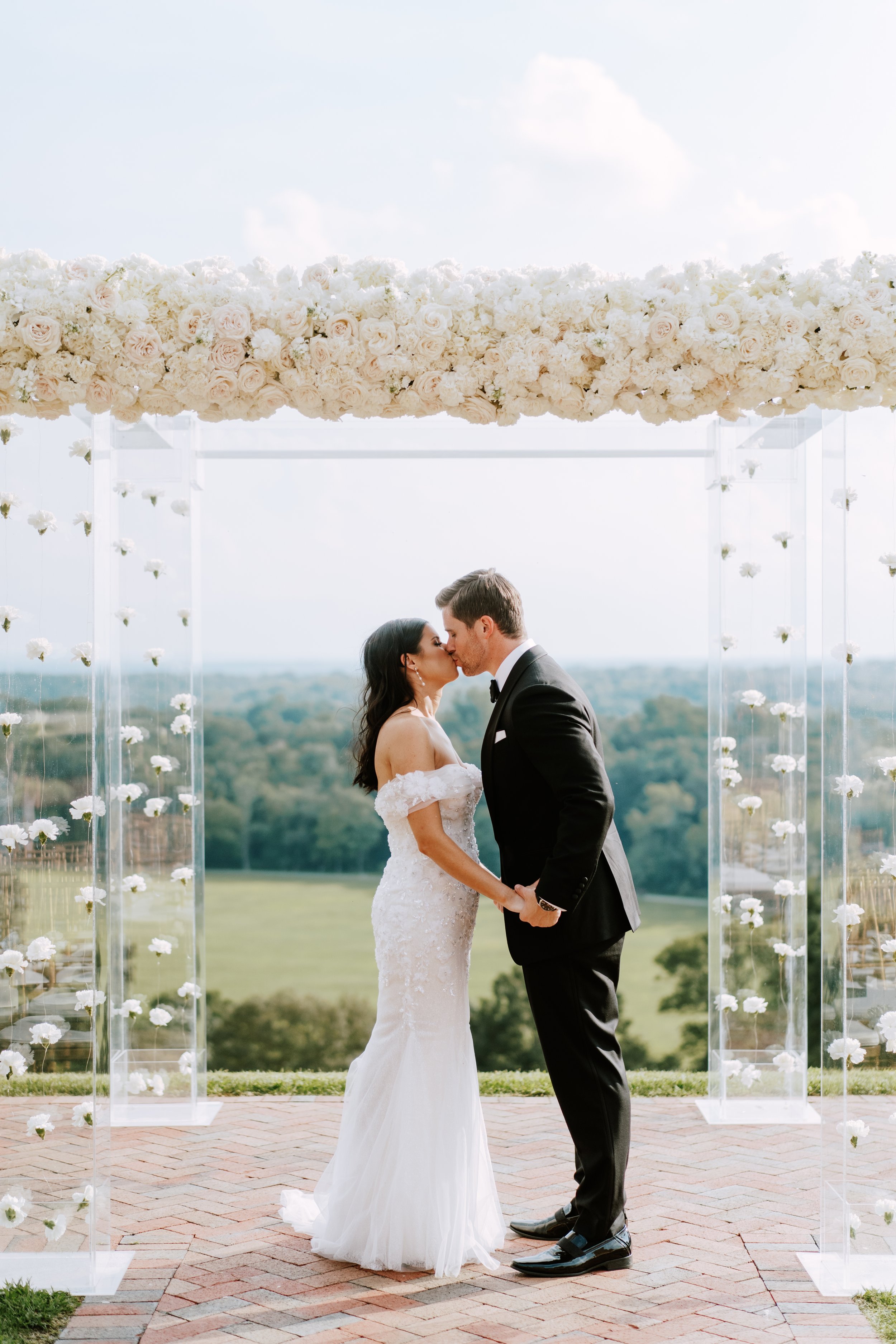 A bride and groom sharing a kiss during their outdoor wedding ceremony under a floral arch with a scenic landscape in the background.