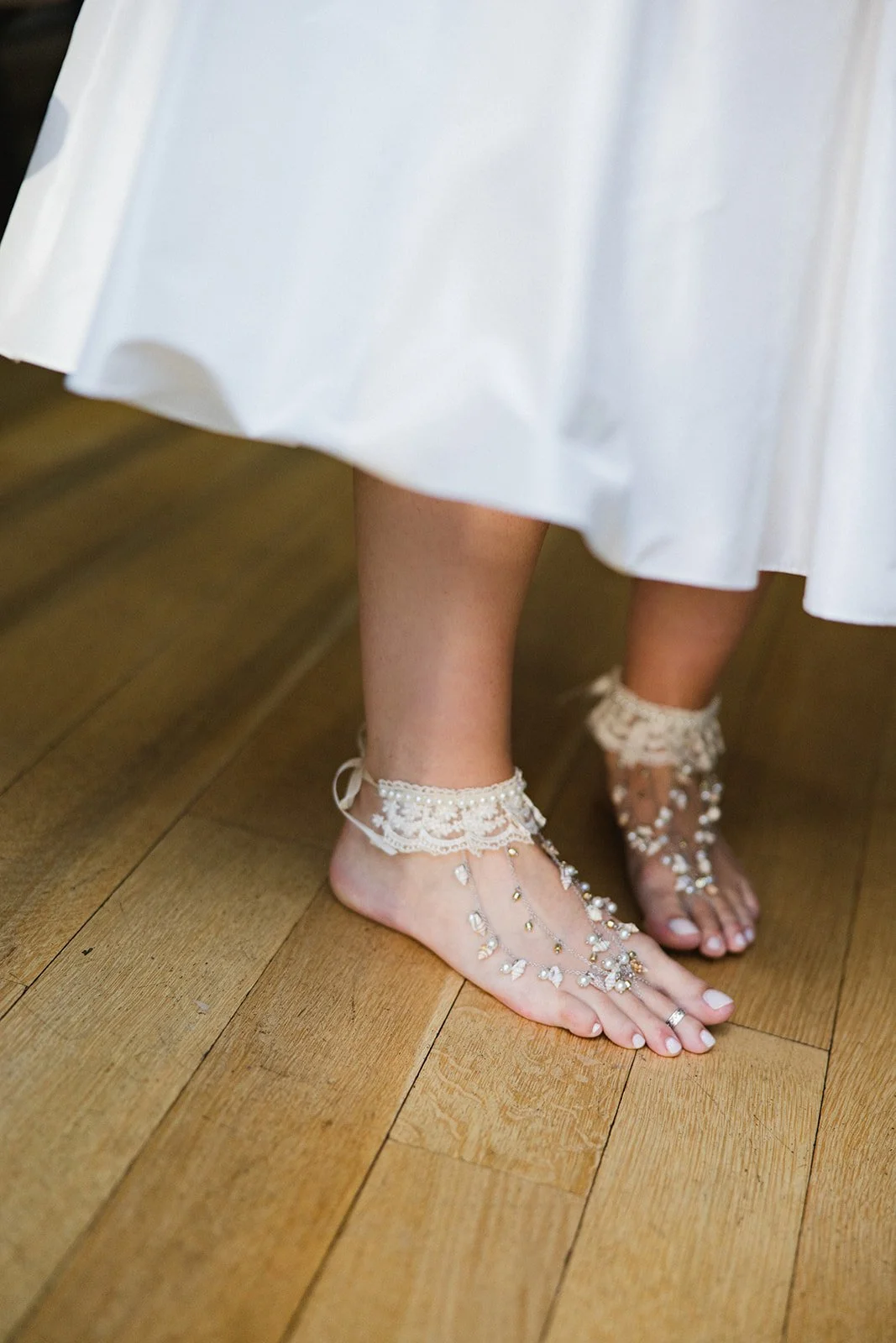 Close-up of a person's bare feet with elaborate pearl and rhinestone hand jewelry, wearing lace anklets, standing on a wooden floor near a white dress.