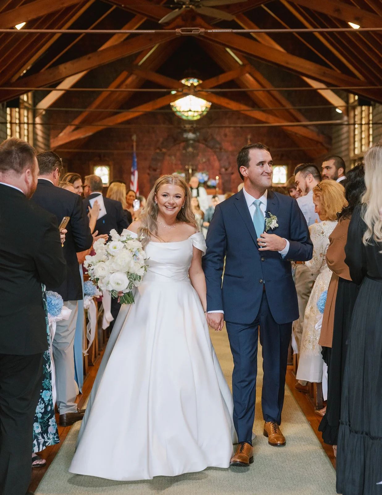 A bride and groom walking down the aisle in a church, smiling, with guests on both sides watching and taking photos.
