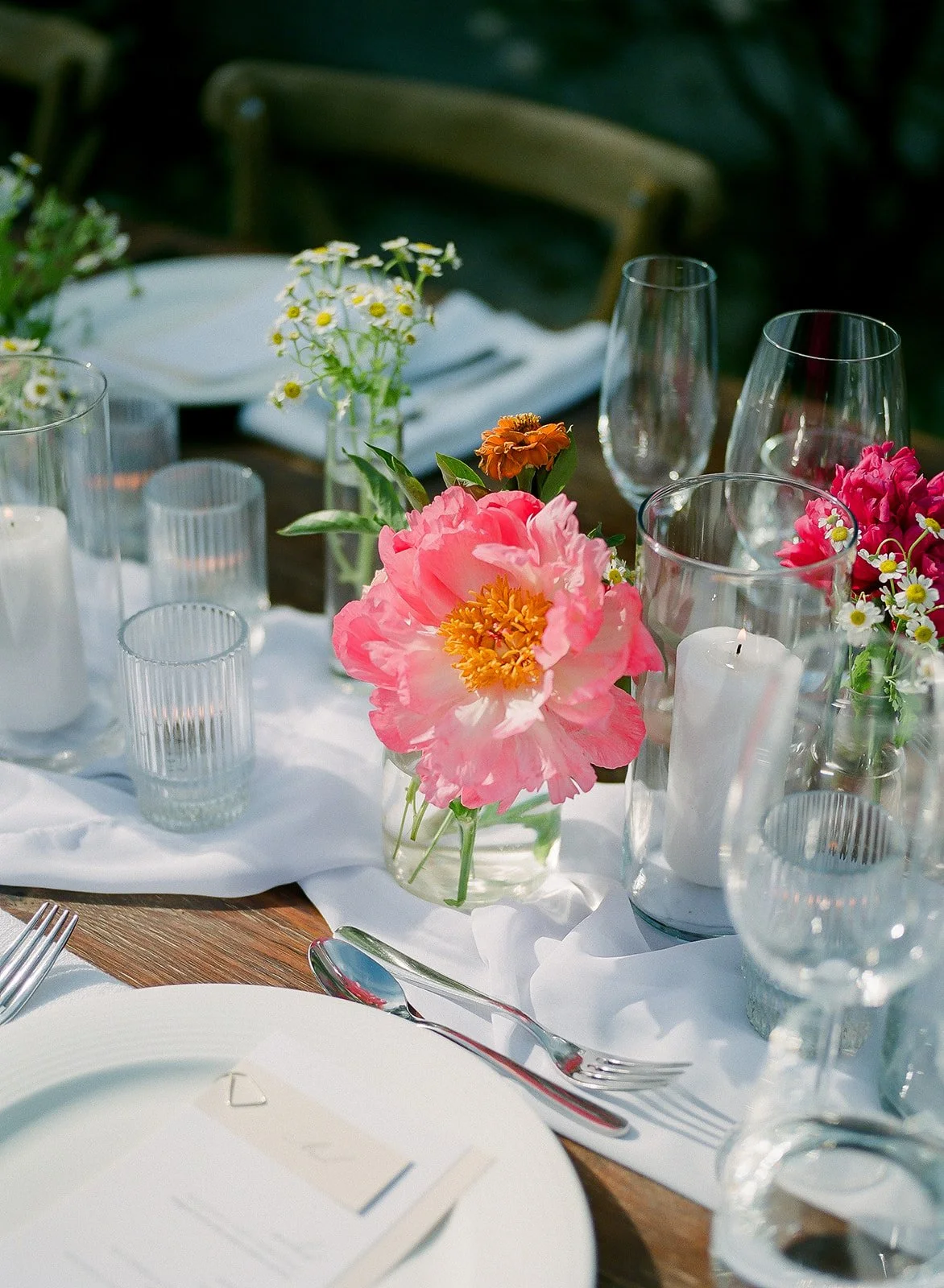 A table decorated with pink and white flowers in glass vases, candles, clear glassware, and white plates, set for an outdoor meal.