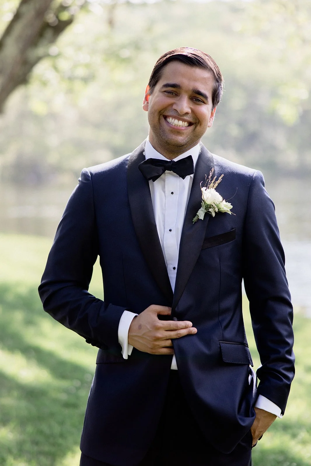 A smiling man in a black tuxedo with a bow tie and a boutonniere stands outdoors with trees and greenery in the background.