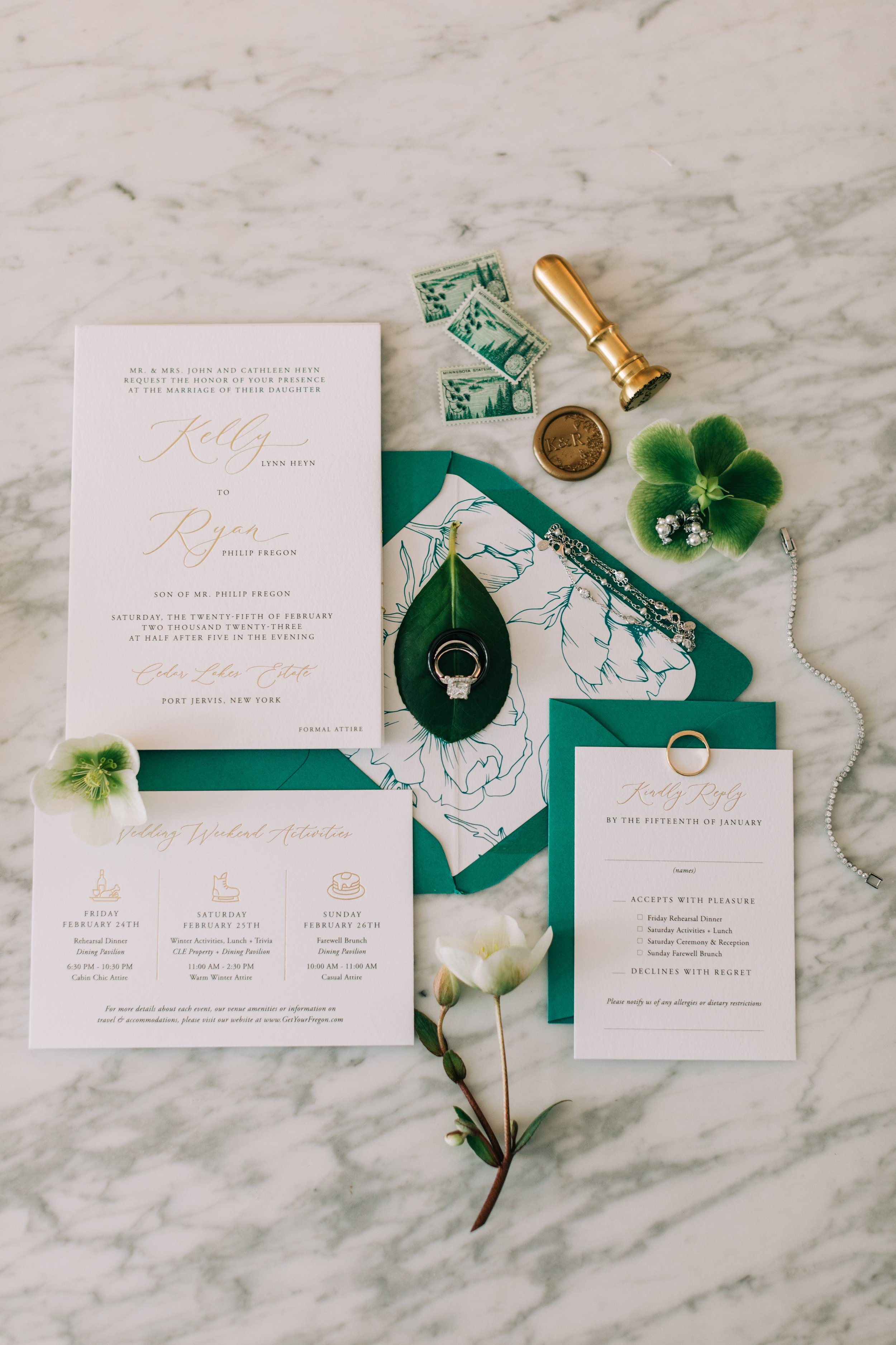 Wedding invitation suite with RSVP card, itinerary card, teal envelopes, floral decorations, jewelry, stamps, wax seal, and a small flower on a marble surface.