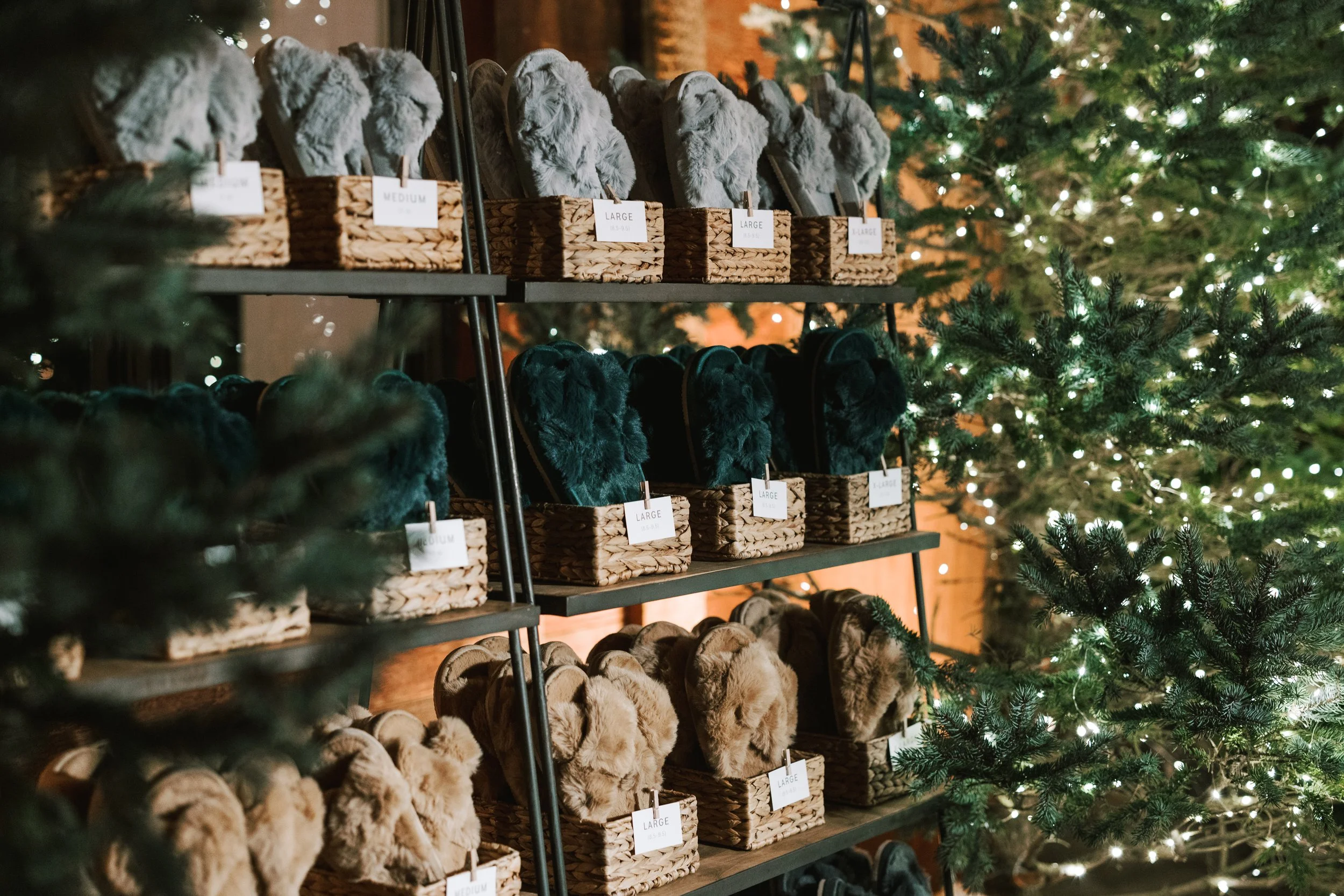 A display of fuzzy slippers in various neutral colors on wooden shelves decorated with baskets, with Christmas trees with lights in the background.