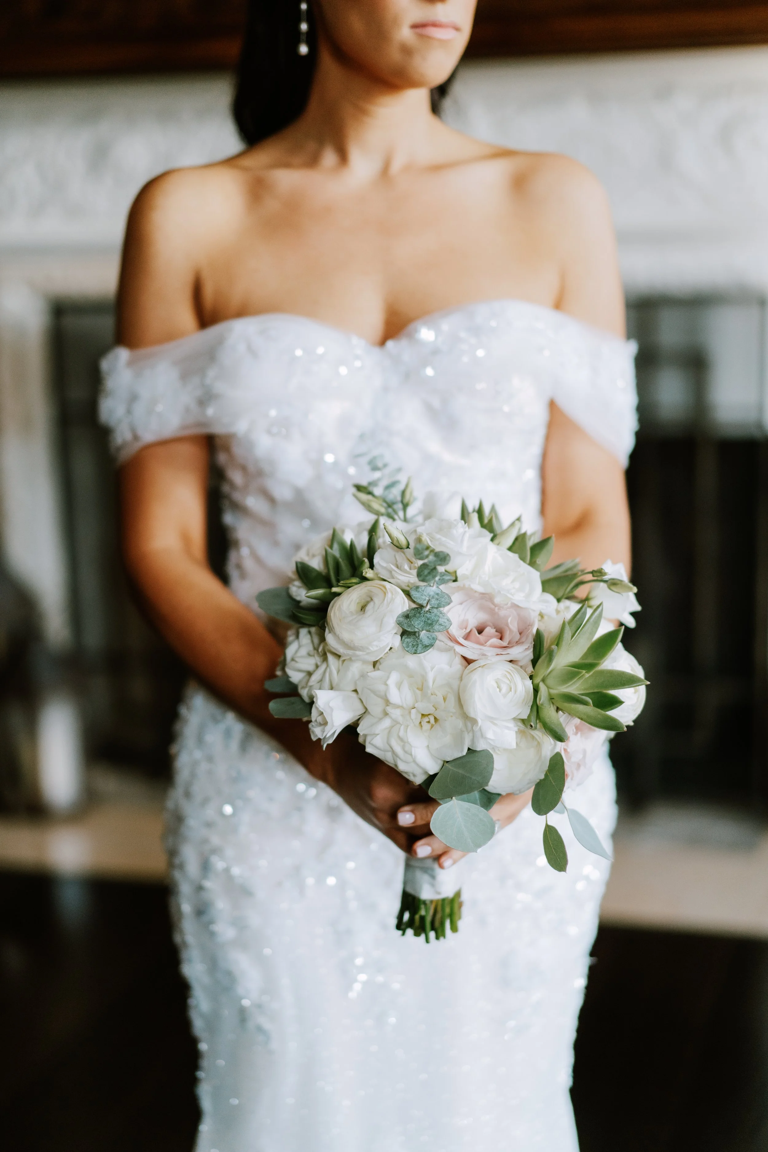 A bride in a white wedding gown holding a bouquet of white and blush pink flowers with green foliage.