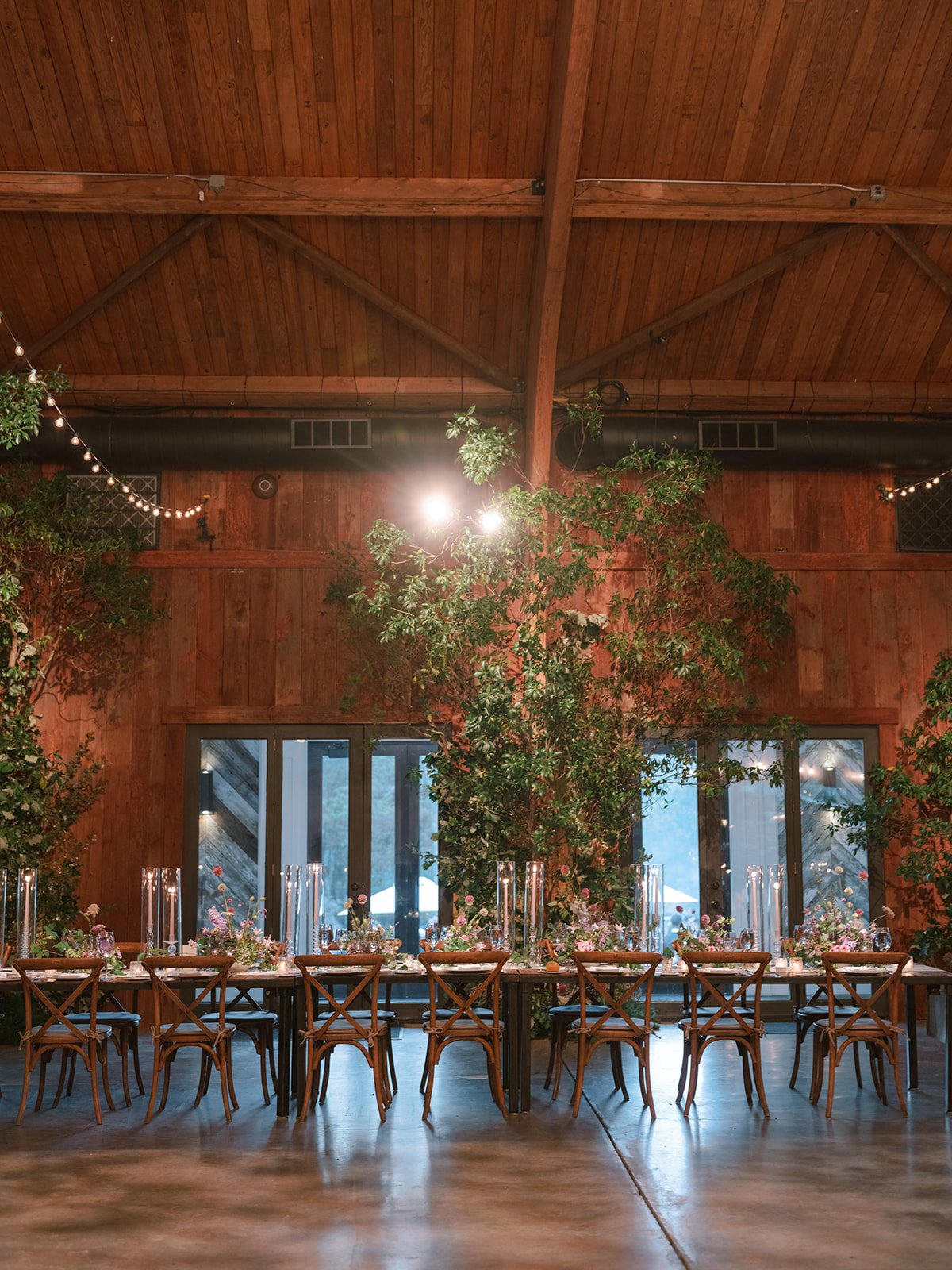 A long banquet table set with floral centerpieces and tall glass candle holders in a rustic indoor venue with wooden walls, large windows, and string lights.
