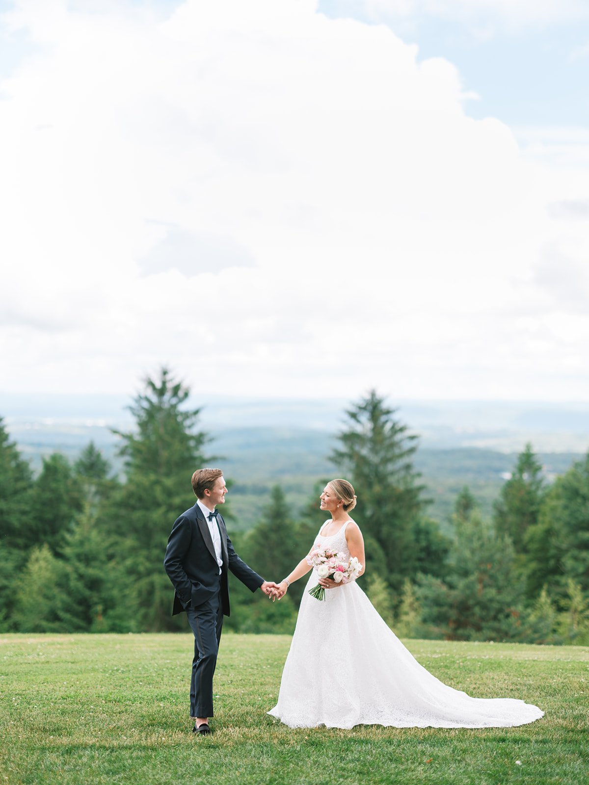 A bride and groom holding hands outdoors on a grassy field with a background of trees and a cloudy sky.