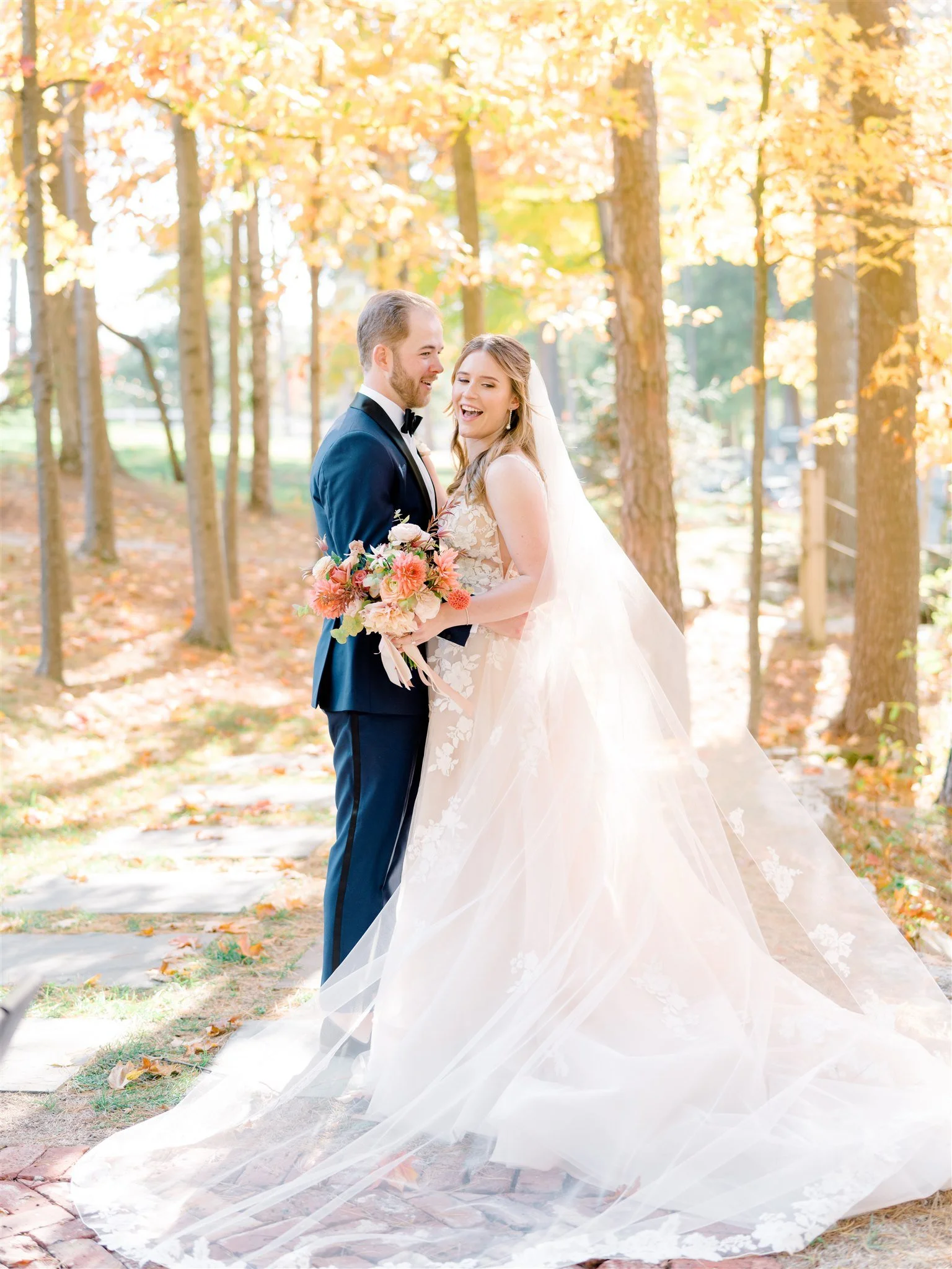 A bride and groom smiling and embracing outdoors surrounded by autumn-colored trees, with the bride holding a bouquet of pink and orange flowers.