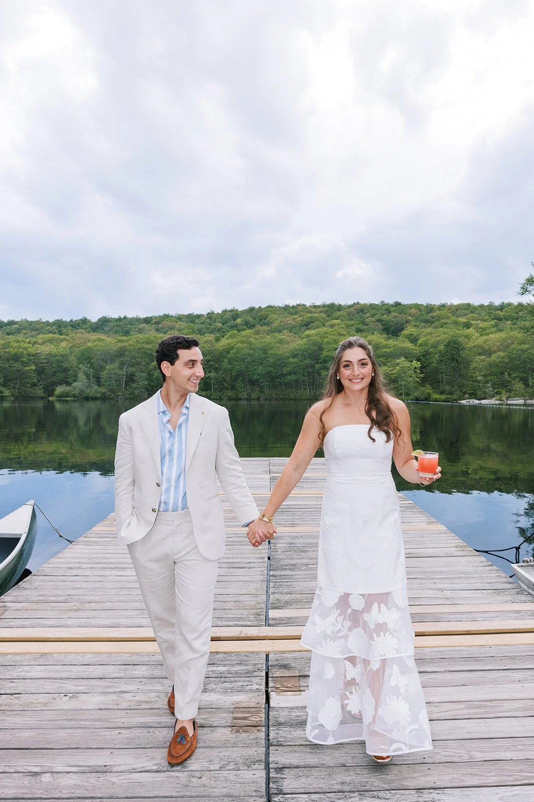 A couple holding hands on a wooden dock by a lake, the woman in a white dress holding a drink, the man in a light suit, with green trees and cloudy sky in the background.