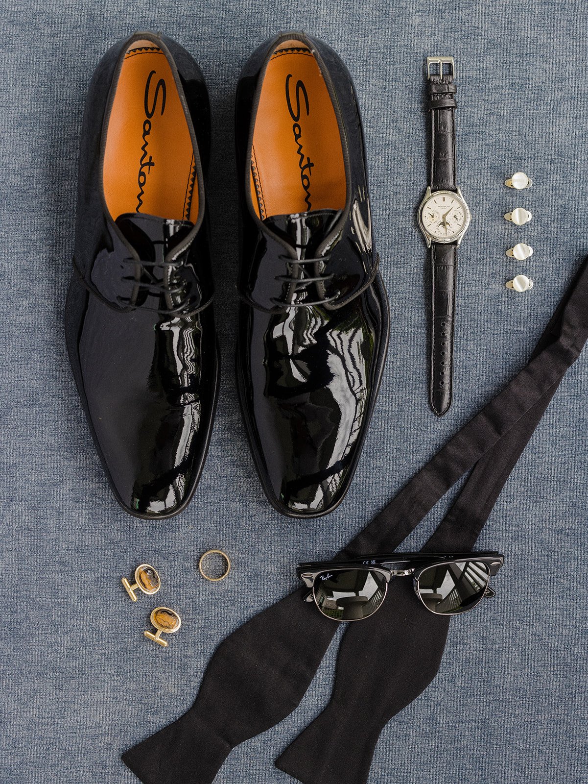 Black shiny dress shoes with orange insoles labeled 'Santon', a black leather watch with a white face, three pairs of pearl earrings, a gold ring, a pair of sunglasses, black socks, and a black tie on a gray fabric surface.
