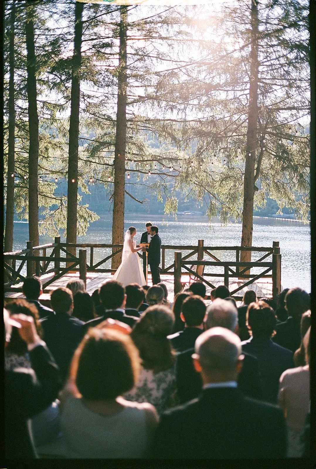 An outdoor wedding ceremony takes place on a wooden platform by a lake surrounded by tall trees. The bride and groom exchange vows, with the officiant standing between them. Guests sit in chairs facing the couple, with sunlight filtering through the 