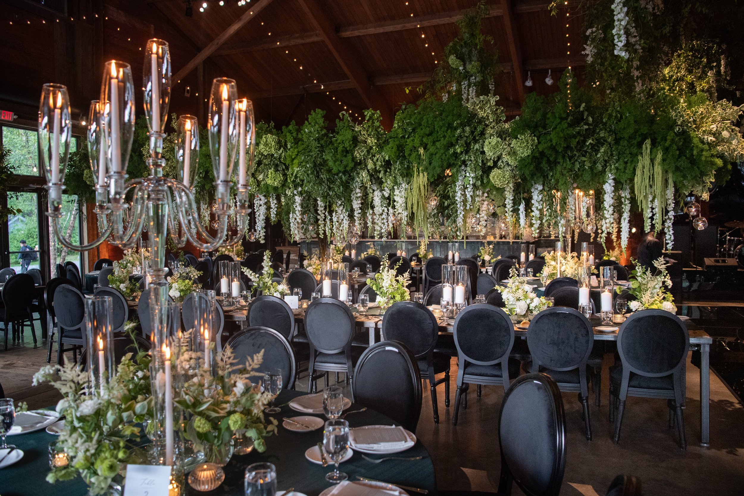 Elegant banquet hall decorated with lush green and white floral arrangements hanging from the ceiling, surrounded by black velvet chairs and tables set with candles, glassware, and floral centerpieces.