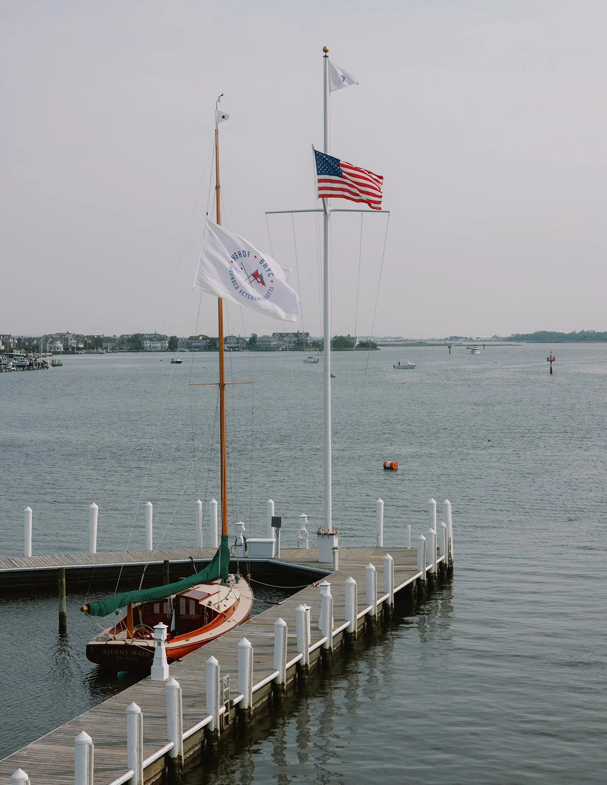 A dock with a small sailboat named Silent Maid moored, and three flags flying on a pole near the water, including the American flag.