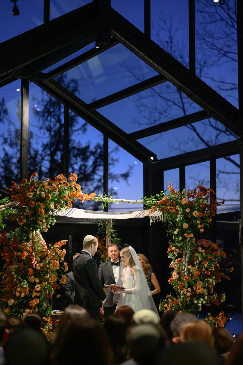 A wedding ceremony taking place indoors under a glass ceiling at night, with a wedding couple standing before an officiant, surrounded by floral arrangements in warm autumn colors.
